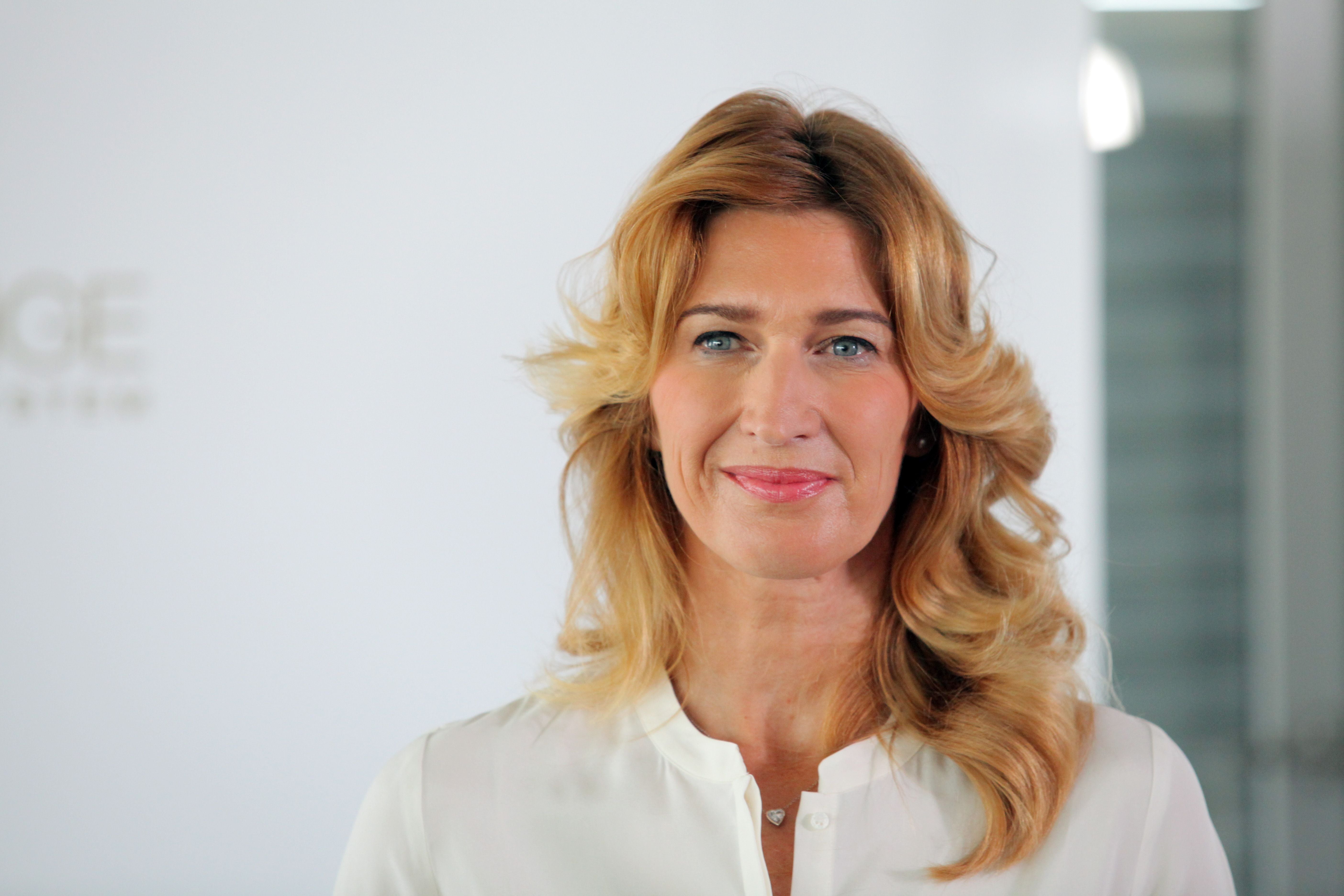 Frau mit blonden, gelockten Haaren und weißer Bluse lächelt vor hellem Hintergrund.