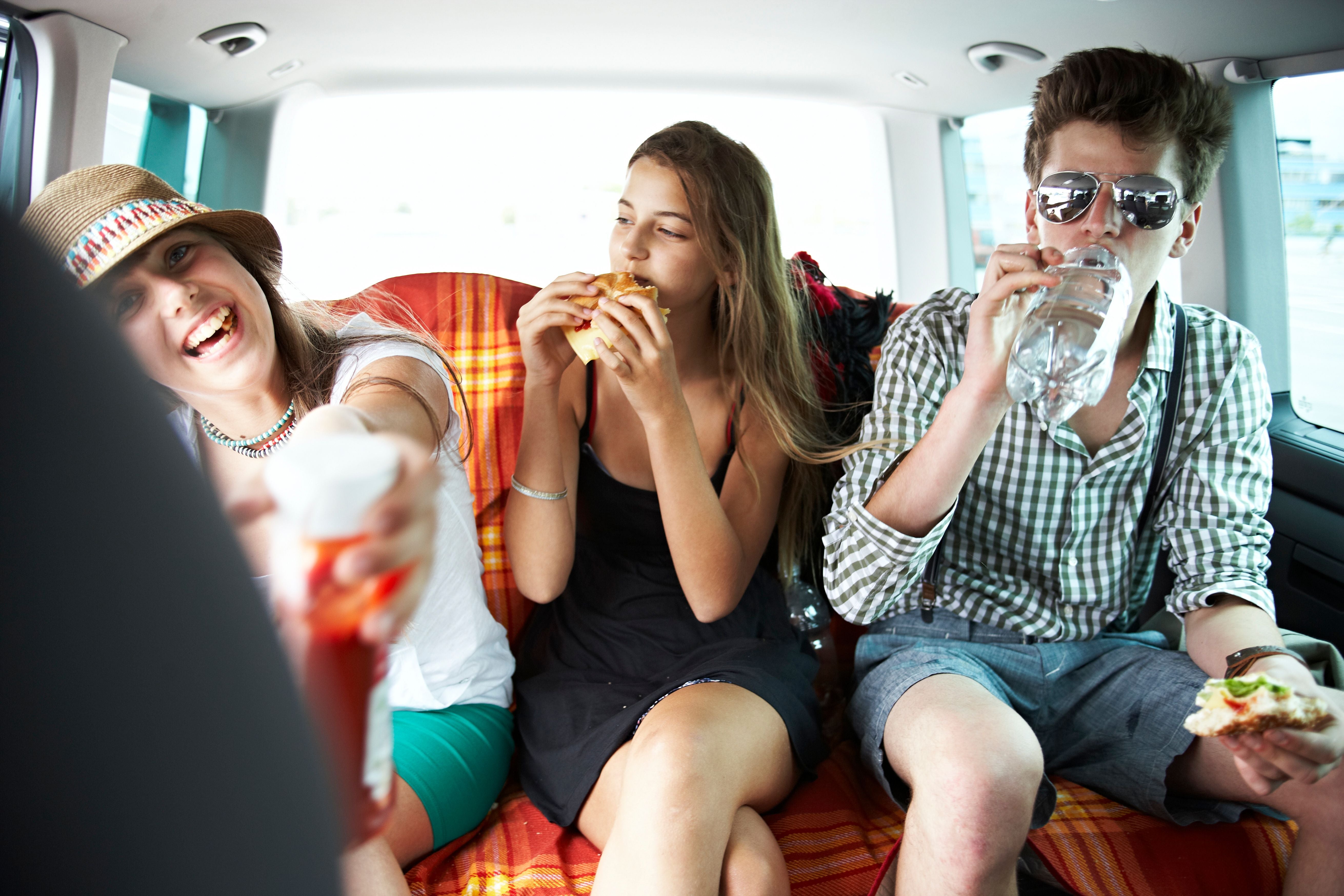 Drei Jugendliche essen und trinken fröhlich auf dem Rücksitz eines Autos.