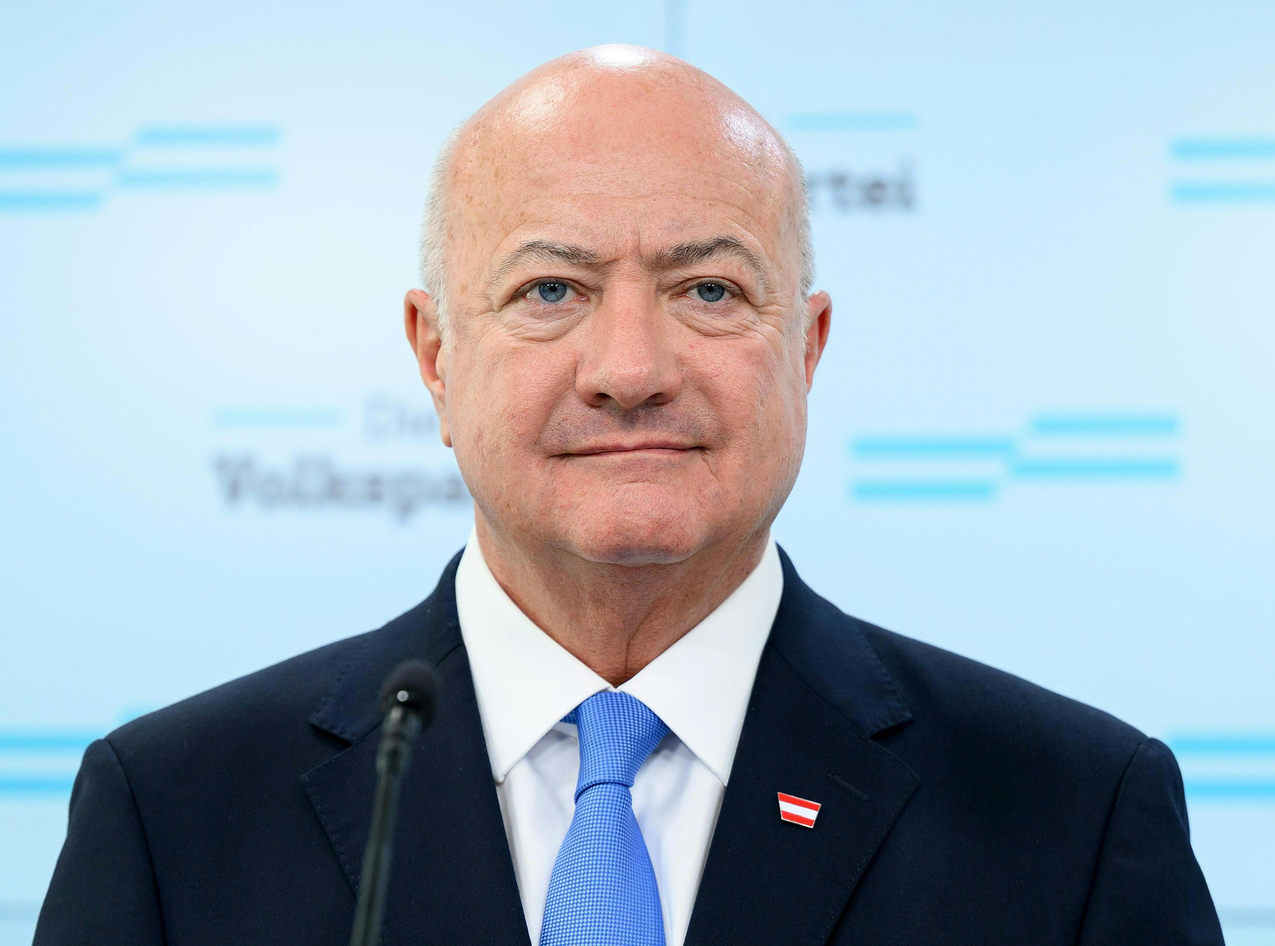 Ein glatzköpfiger Mann im Anzug steht vor einem Mikrofon bei einer Pressekonferenz.