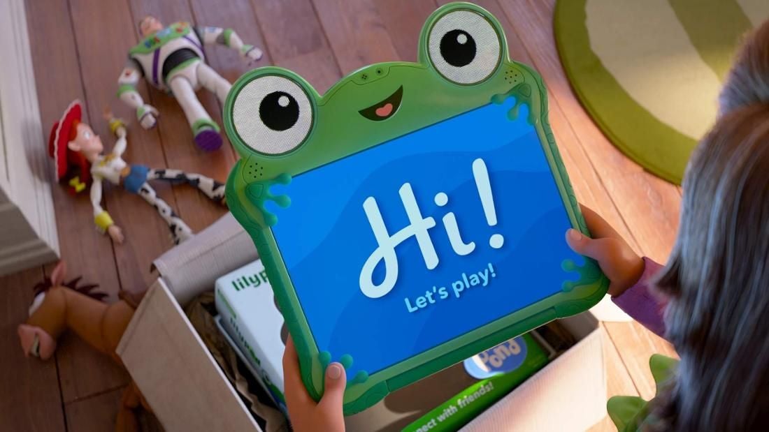 Ein Kind hält ein Frosch-Tablet mit "Hi! Let's play!" während Toy-Story-Figuren auf dem Boden liegen.