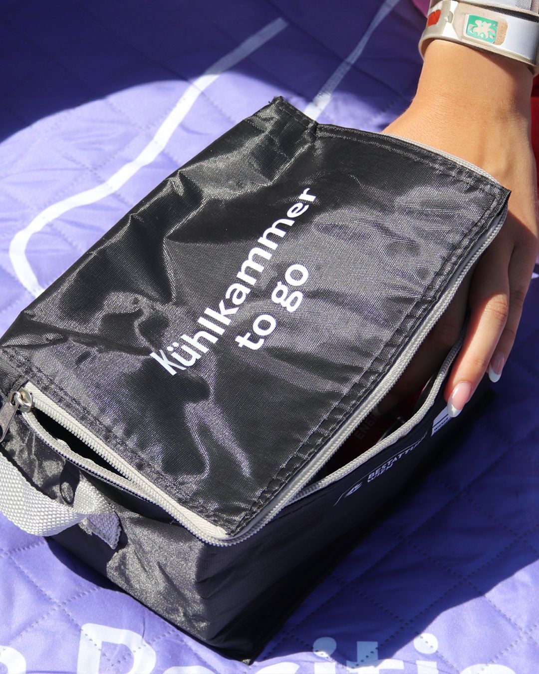 Schwarze Kühltasche mit der Aufschrift „Kühlkammer to go“ auf lila Unterlage, eine Hand hält sie fest.