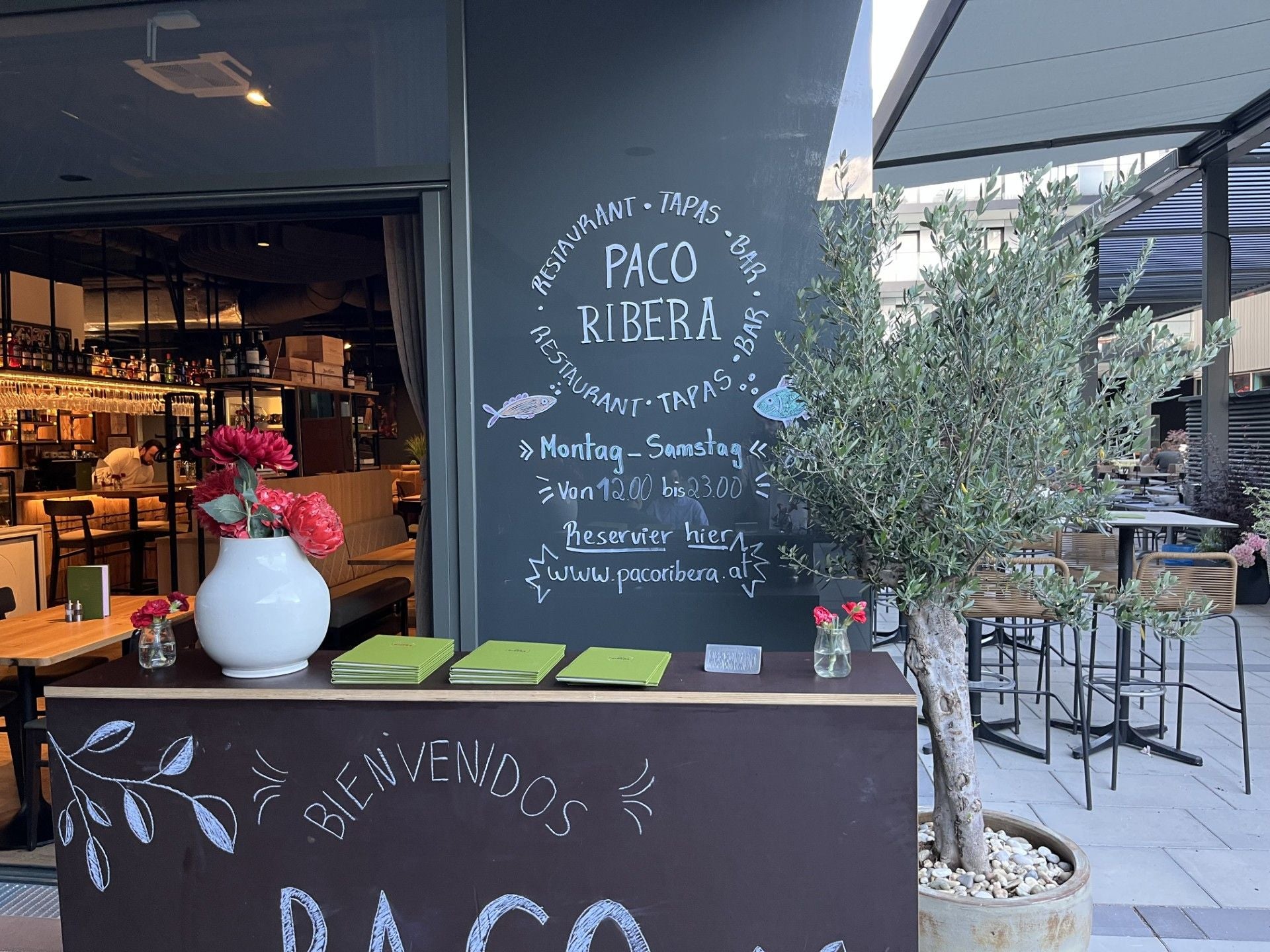Eingang eines Restaurants namens Paco Ribera mit Menüs, Blumenvasen und einer Schiefertafel.