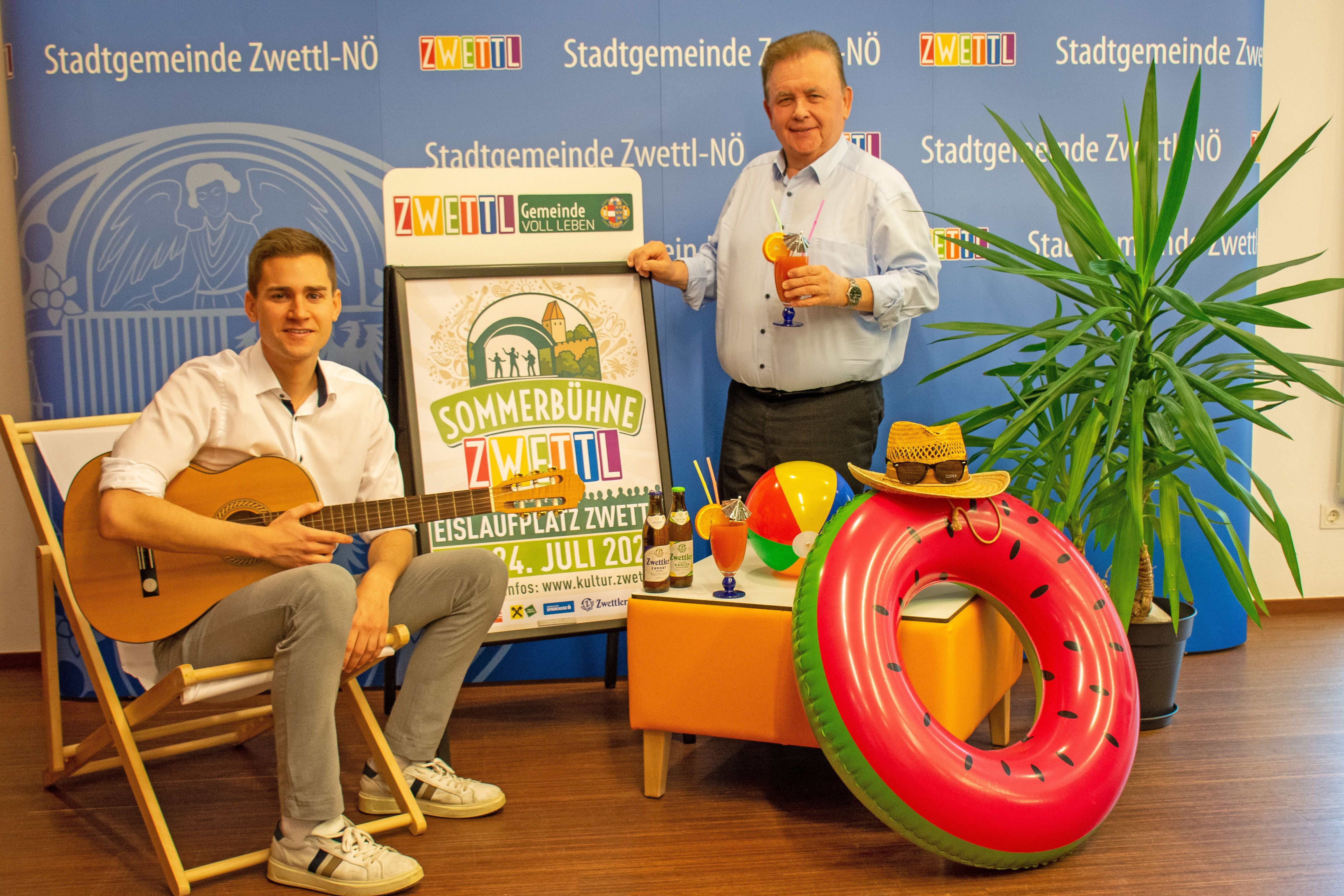 Zwei Männer posieren bei einer Sommerbühne-Zwettl-Werbung mit Gitarre und Sommerdeko.