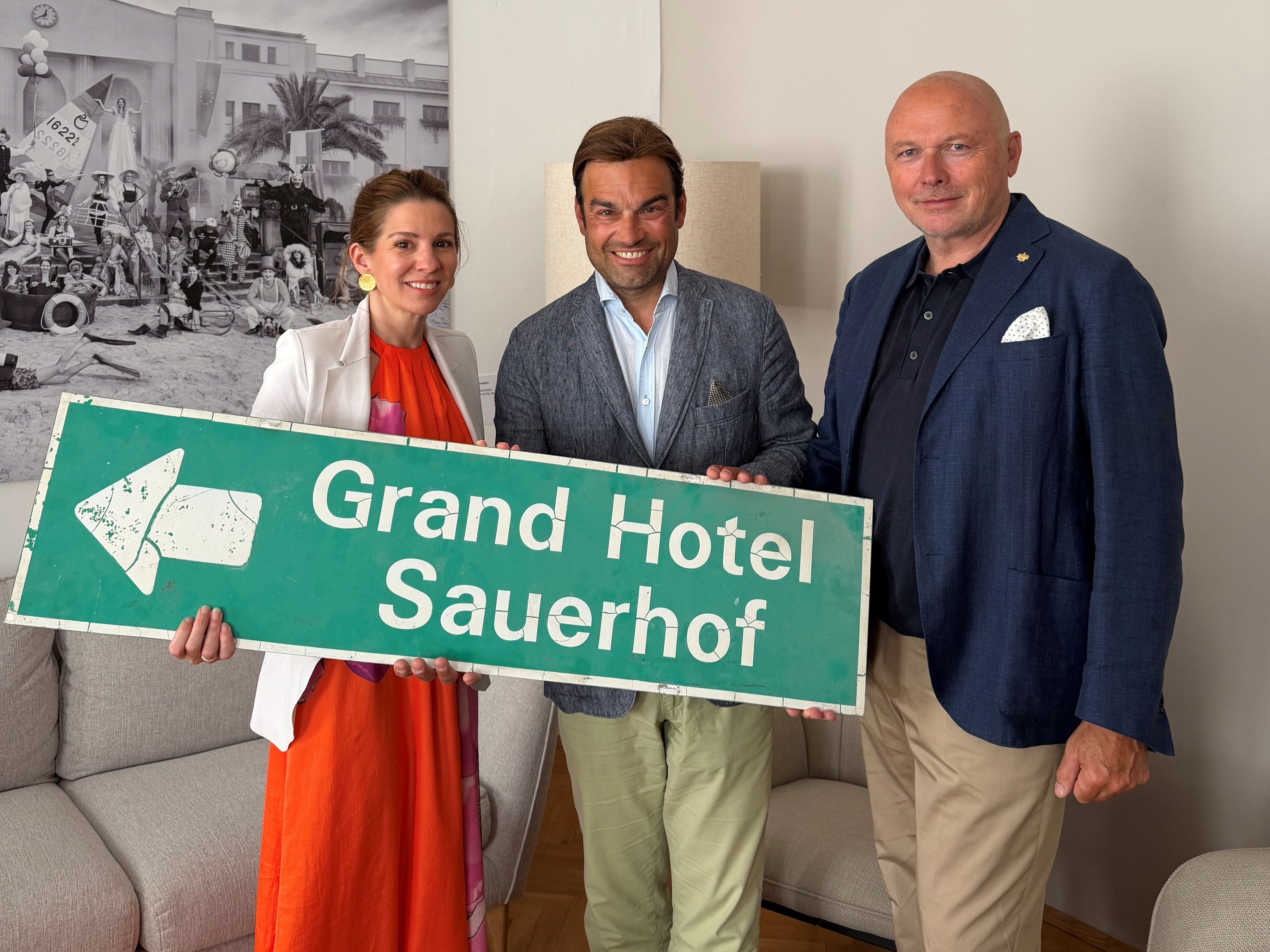 Drei Personen halten ein grünes Schild mit der Aufschrift „Grand Hotel Sauerhof“.