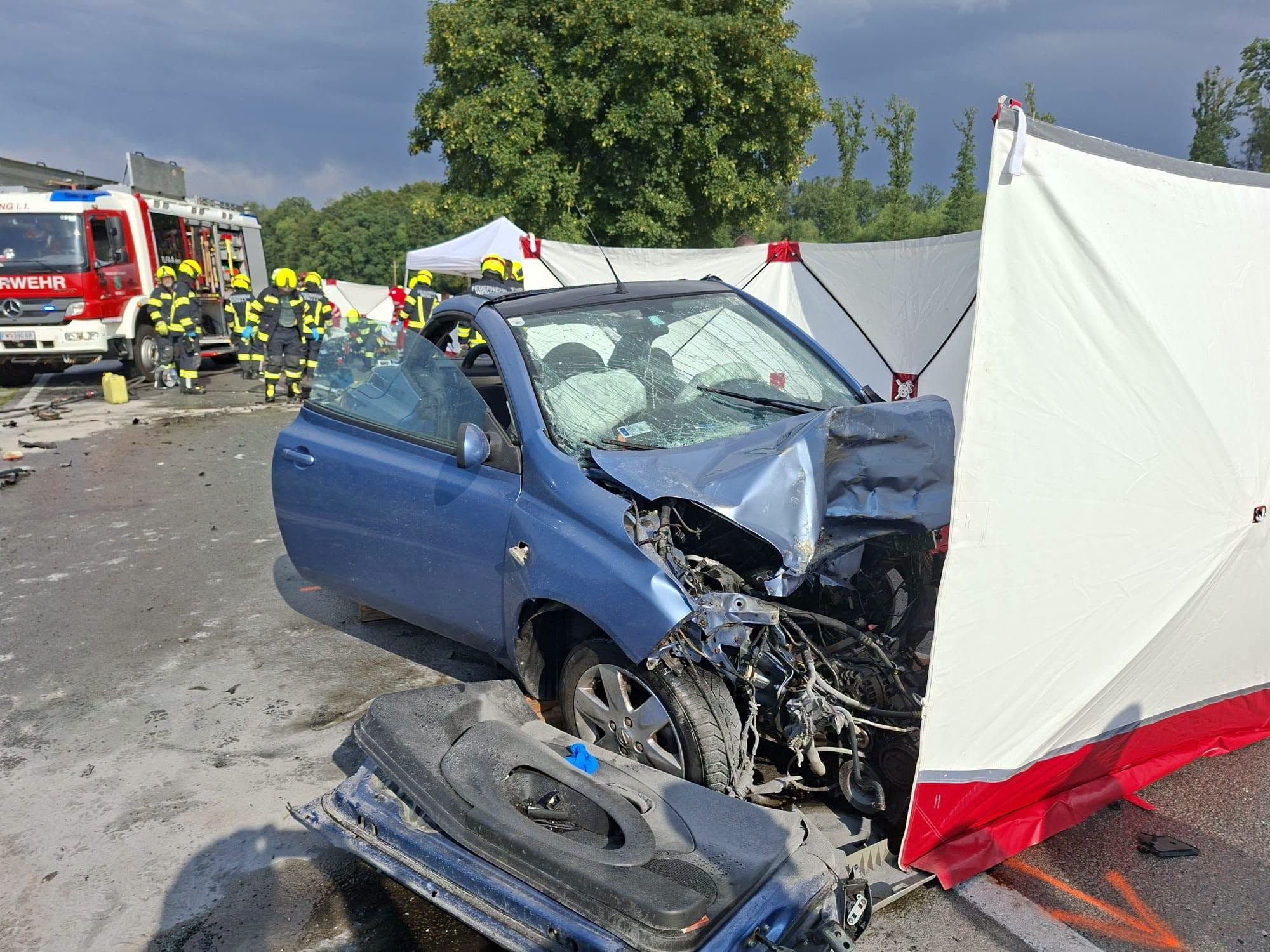 Schwer beschädigtes Auto nach Unfall, Feuerwehrleute und Einsatzfahrzeug im Hintergrund.