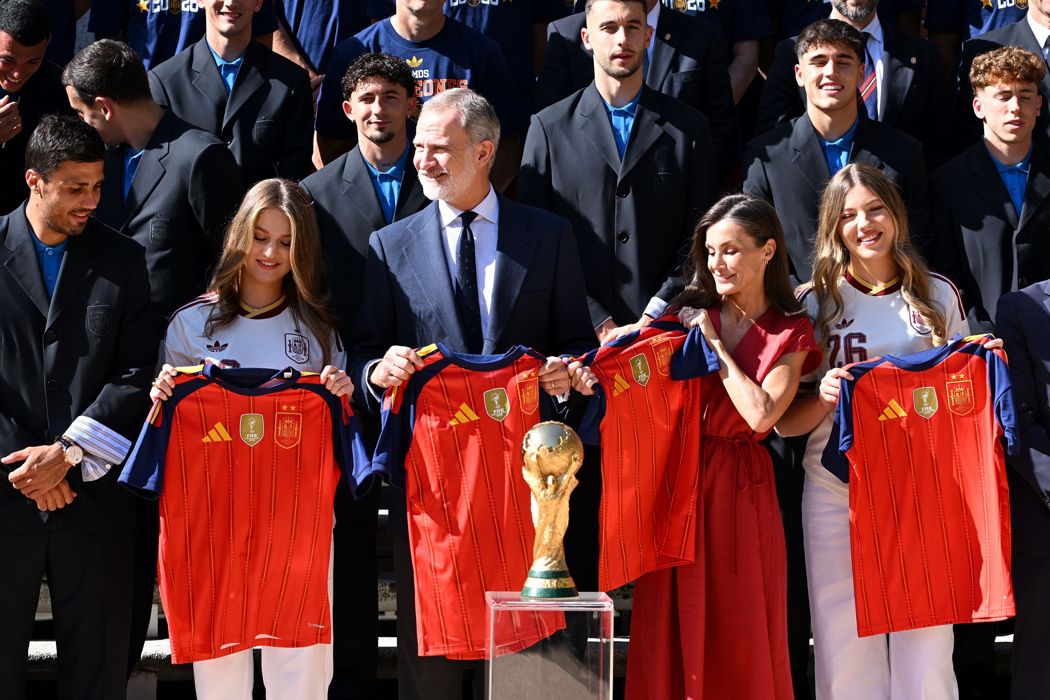 Der spanische König und seine Familie halten Spanien-Trikots vor dem WM-Pokal, umgeben vom Fußballteam.