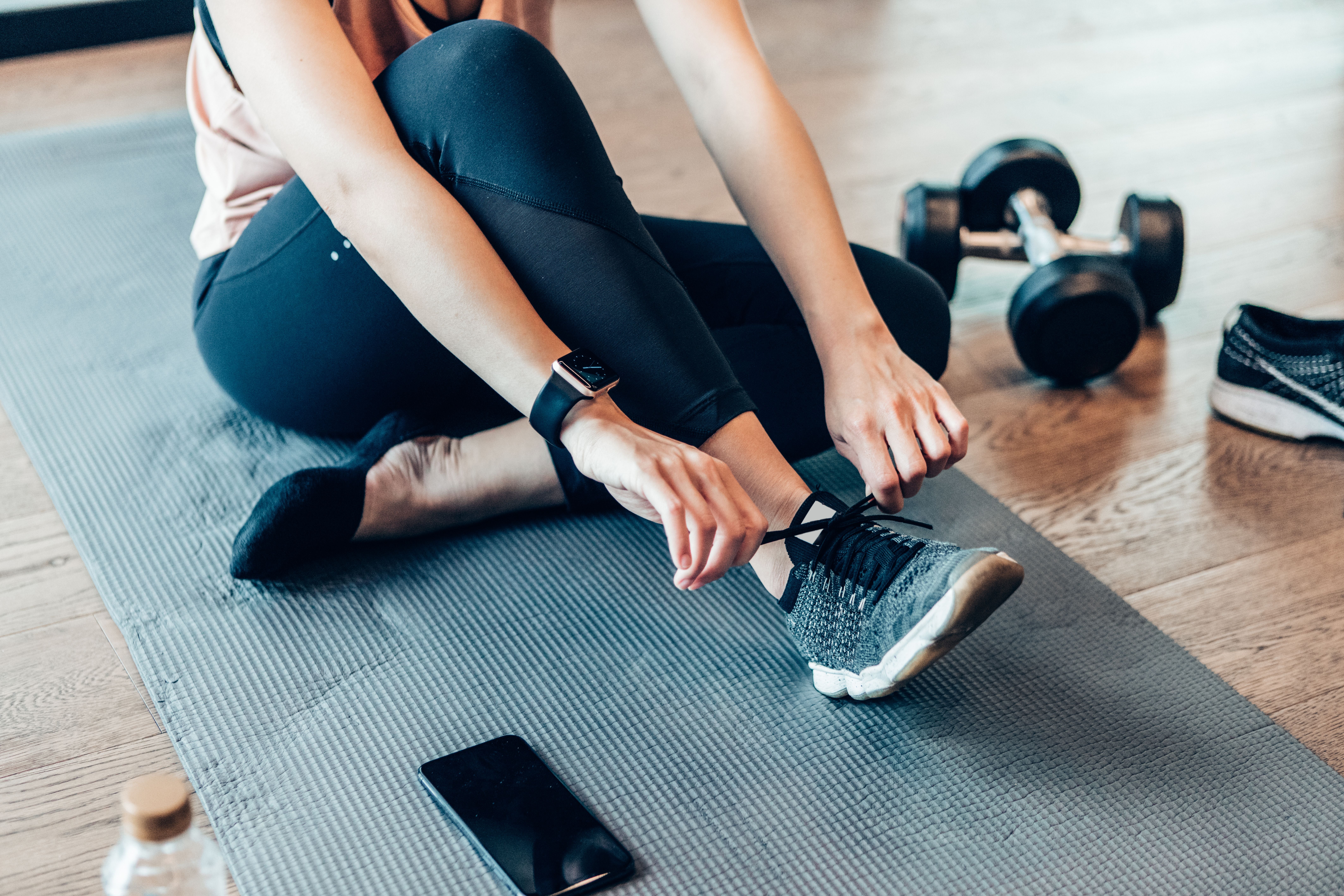 Person bindet Sportschuh auf Yogamatte; daneben liegt ein Handy und eine Hantel.