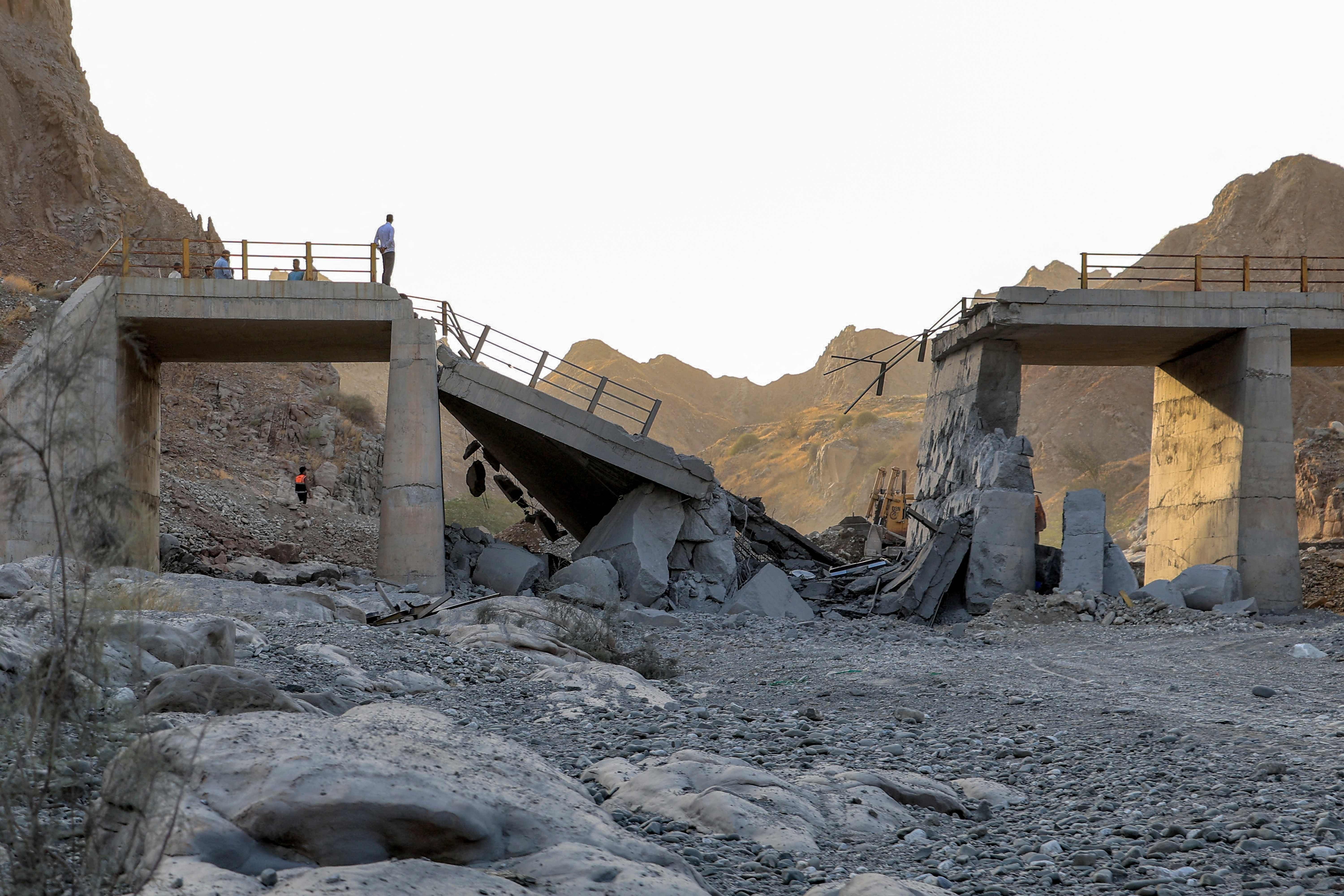 Zerstörte Brücke in bergiger Landschaft, Menschen stehen auf den Bruchstellen der Brücke.