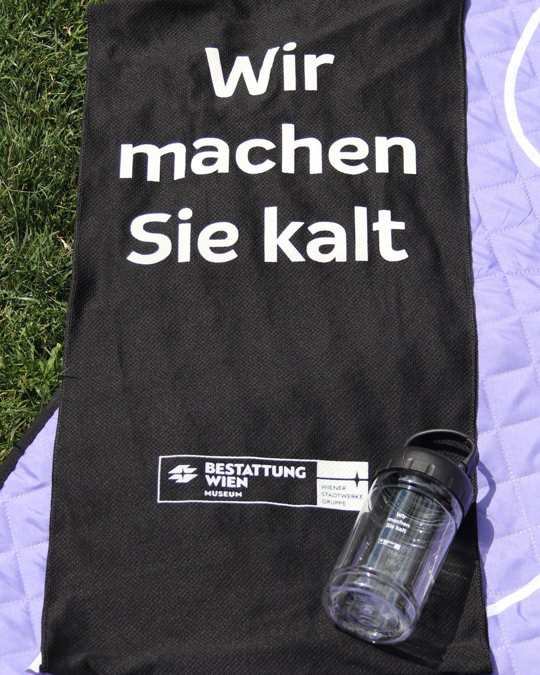 Schwarzes Kühltuch mit Aufdruck „Wir machen Sie kalt“, daneben eine Trinkflasche auf einer Decke.