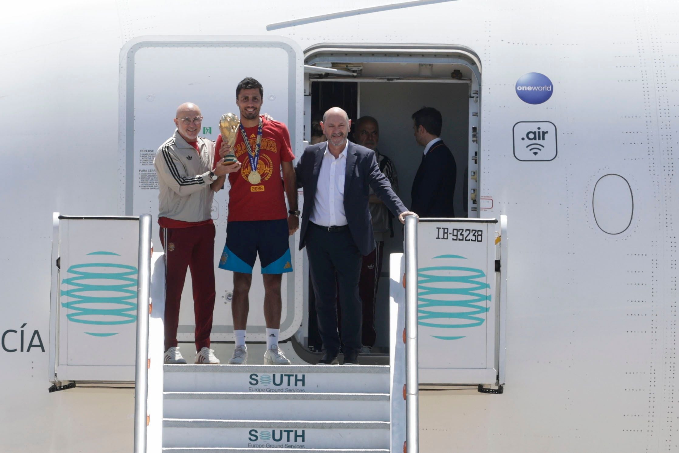 Drei Männer mit WM-Pokal stehen vor einem Flugzeug am Flughafen Madrid nach Spaniens WM-Sieg 2026.
