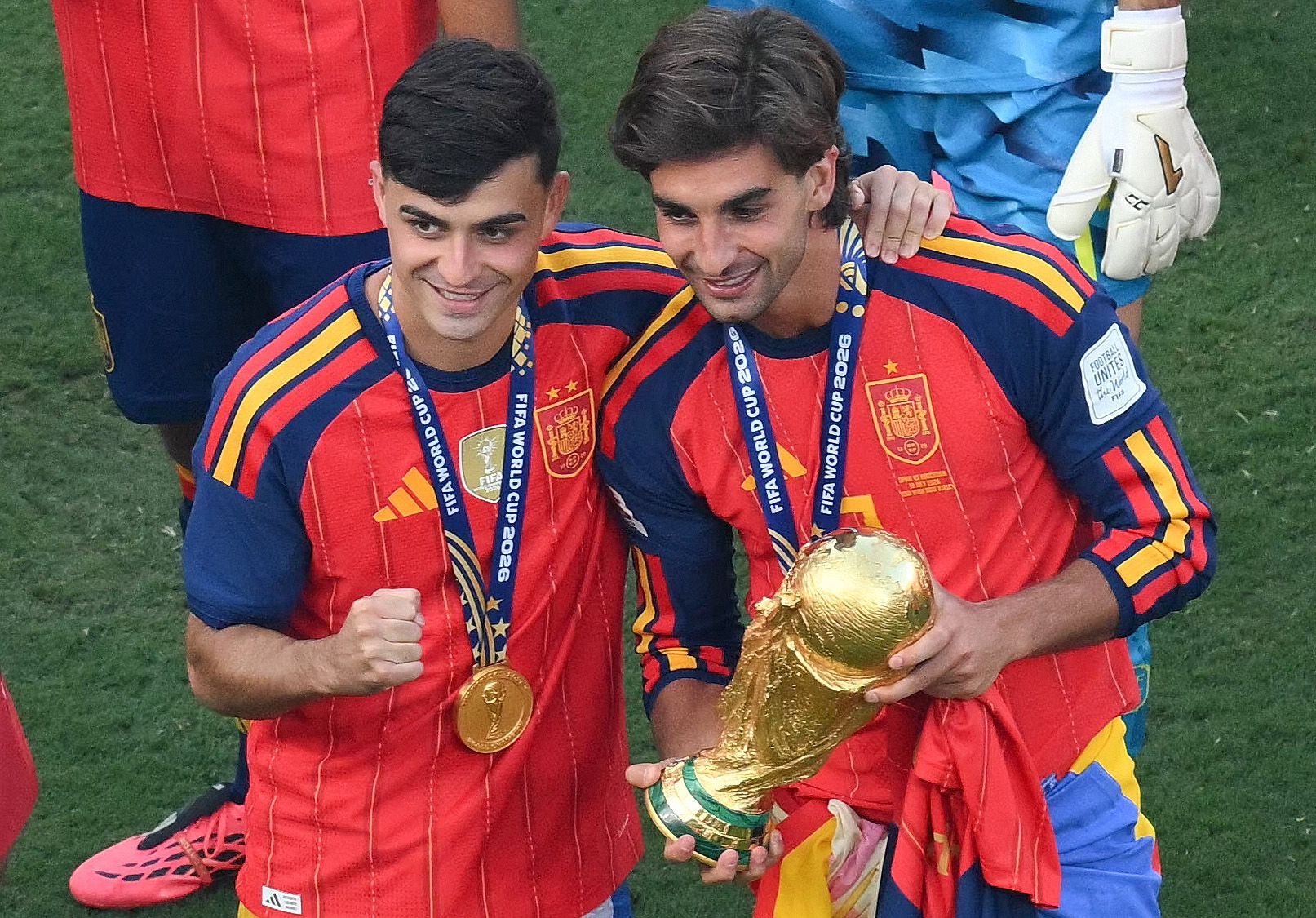 Zwei spanische Fußballspieler feiern mit Medaillen und dem WM-Pokal auf dem Spielfeld.