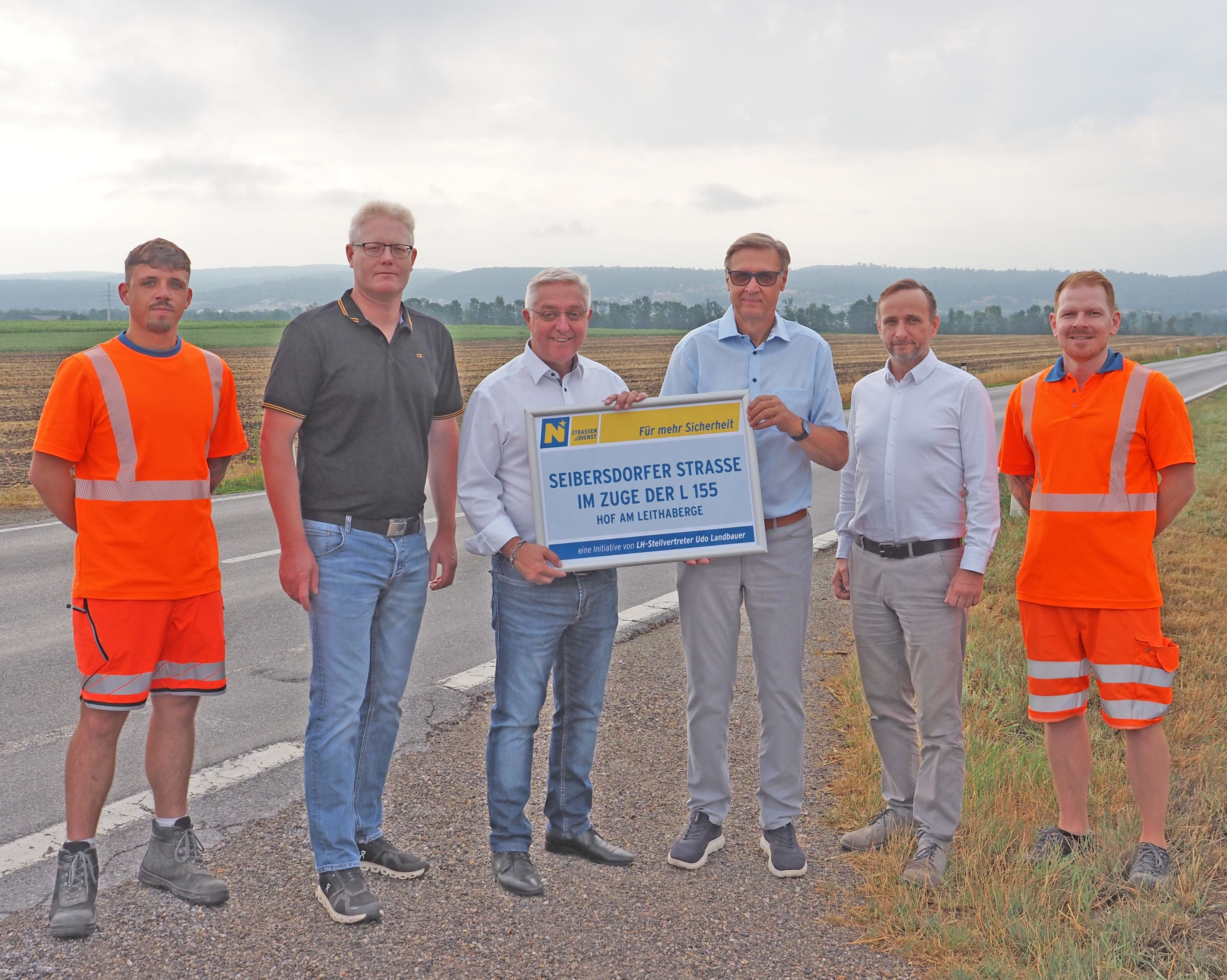 Sechs Männer stehen auf einer Landstraße, zwei davon halten ein Schild zur Straßensicherheit.