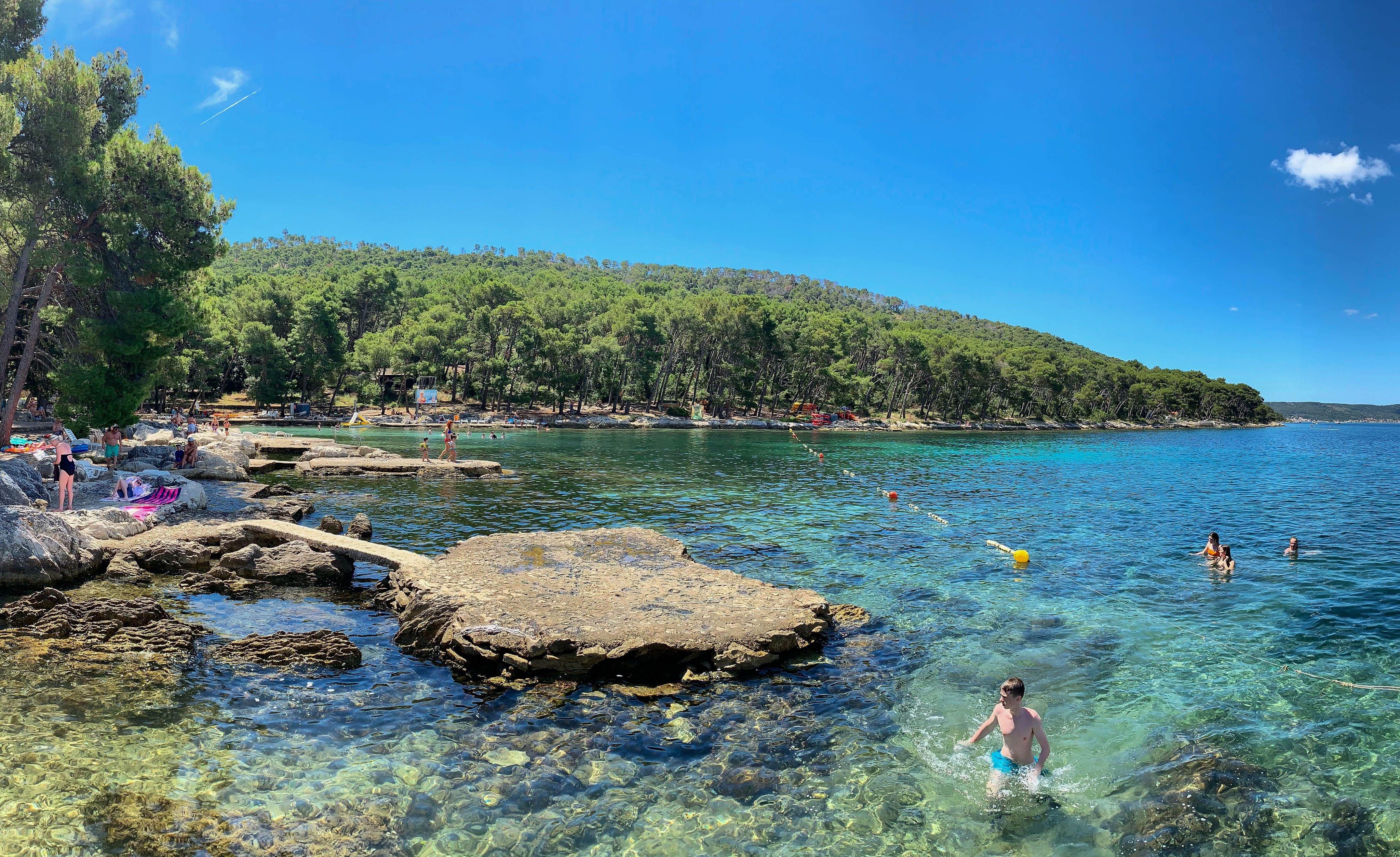 Menschen schwimmen und sonnen sich an einem felsigen Strand mit klarem Wasser und Pinien im Hintergrund.