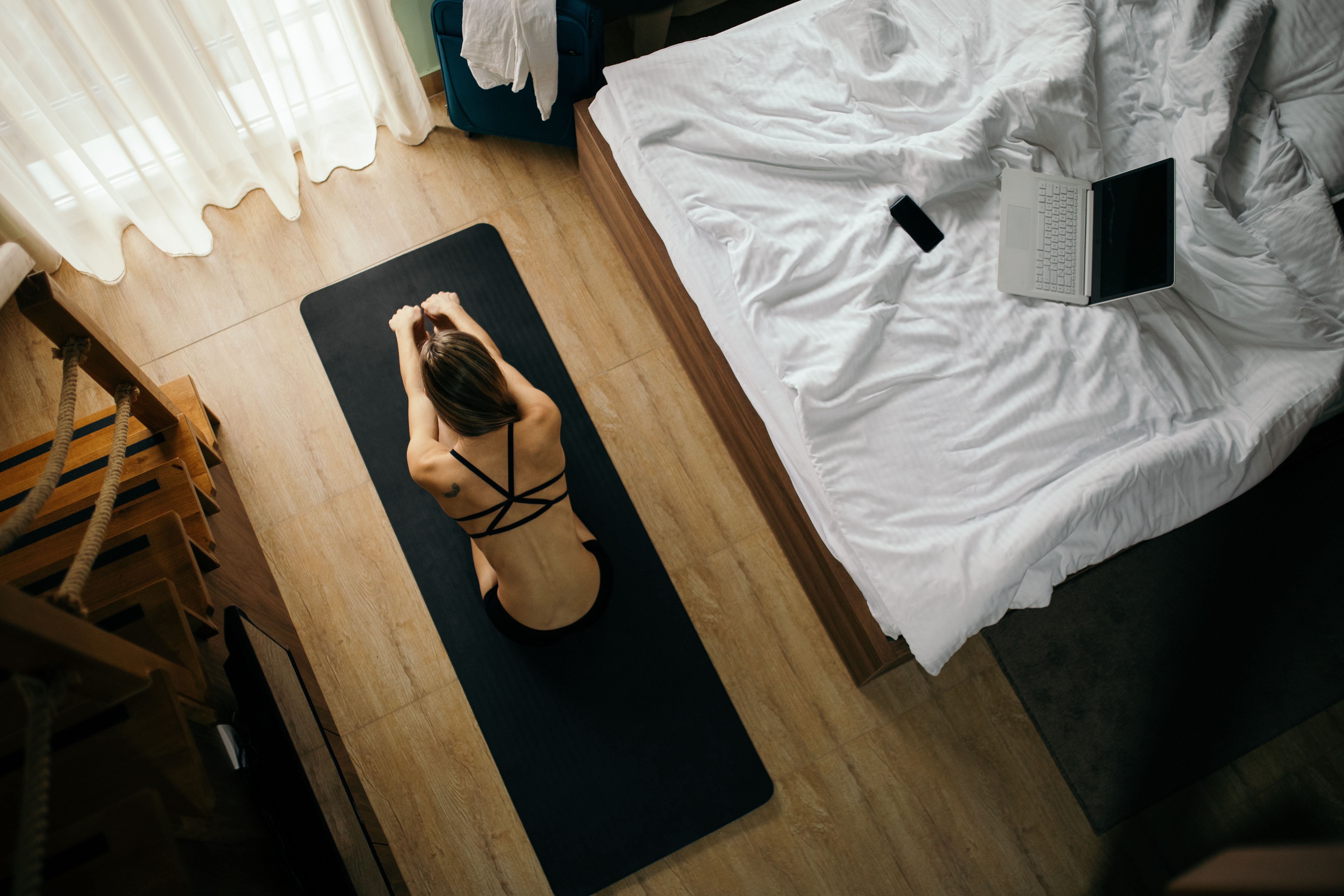 Junge Frau macht Dehnübungen auf einer Yogamatte neben einem ungemachten Bett im Zimmer.