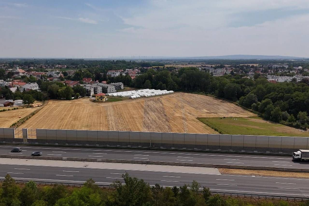 Blick auf ein Feld mit Gebäuden am Stadtrand, davor verläuft eine Autobahn mit Lärmschutzwand.