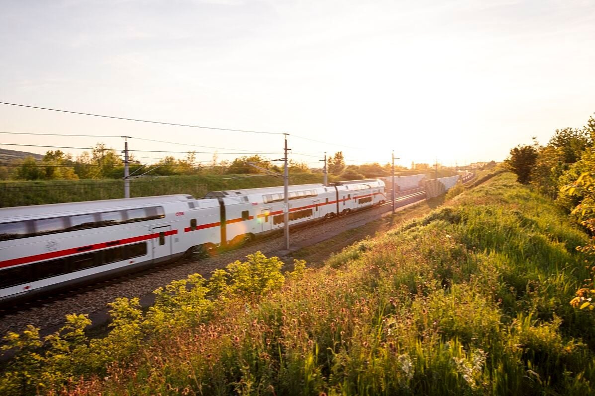 Ein weißer Doppelstockzug fährt bei Sonnenuntergang durch eine schöne grüne Landschaft.