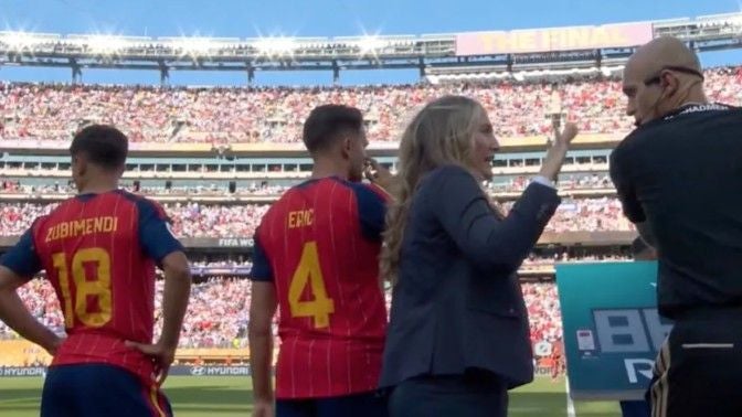 Zwei Fußballspieler im roten Trikot und Offizielle am Spielfeldrand in einem vollen Stadion.