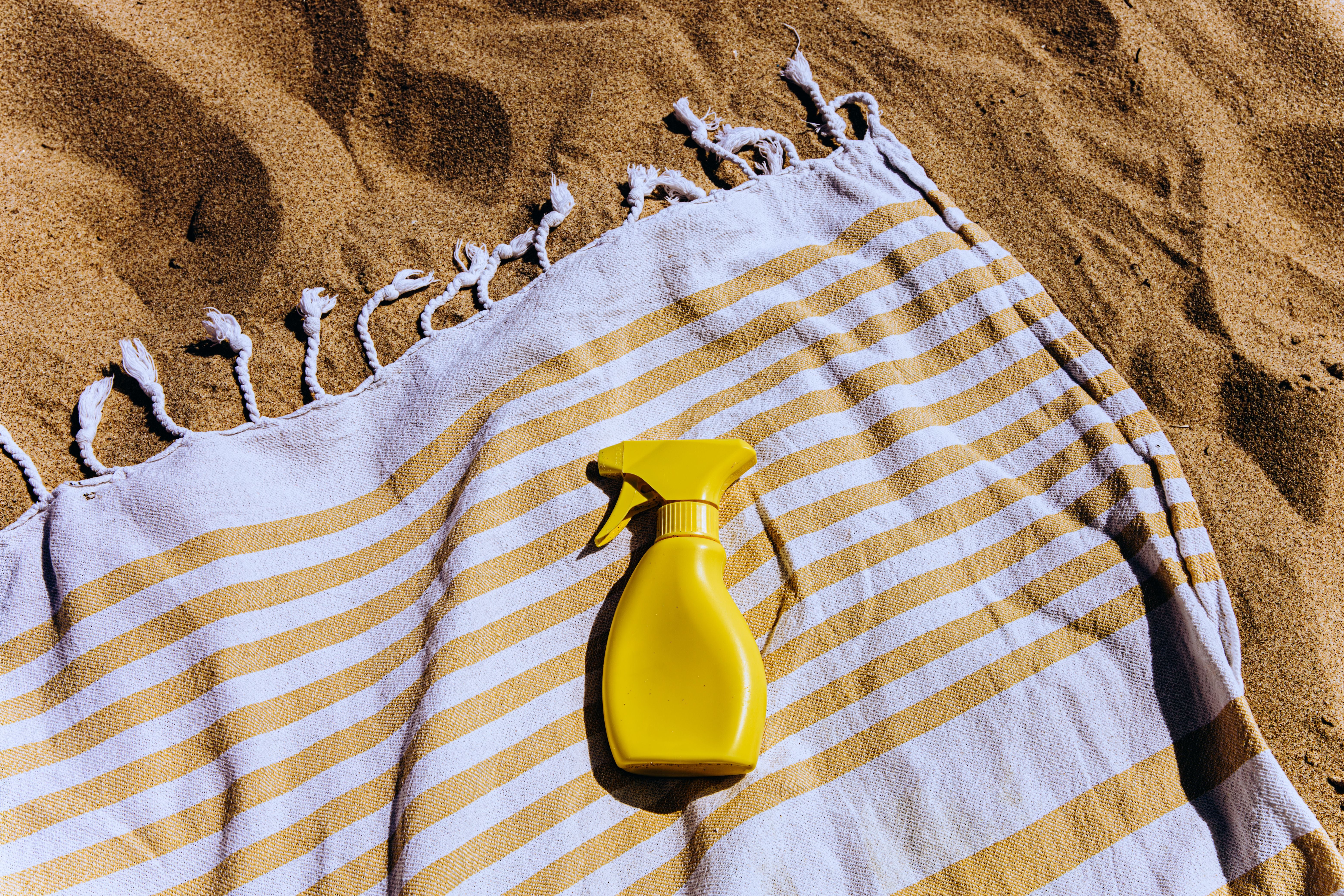 Gelbe Sprühflasche auf einem gelb-weiß gestreiften Handtuch am Sandstrand.