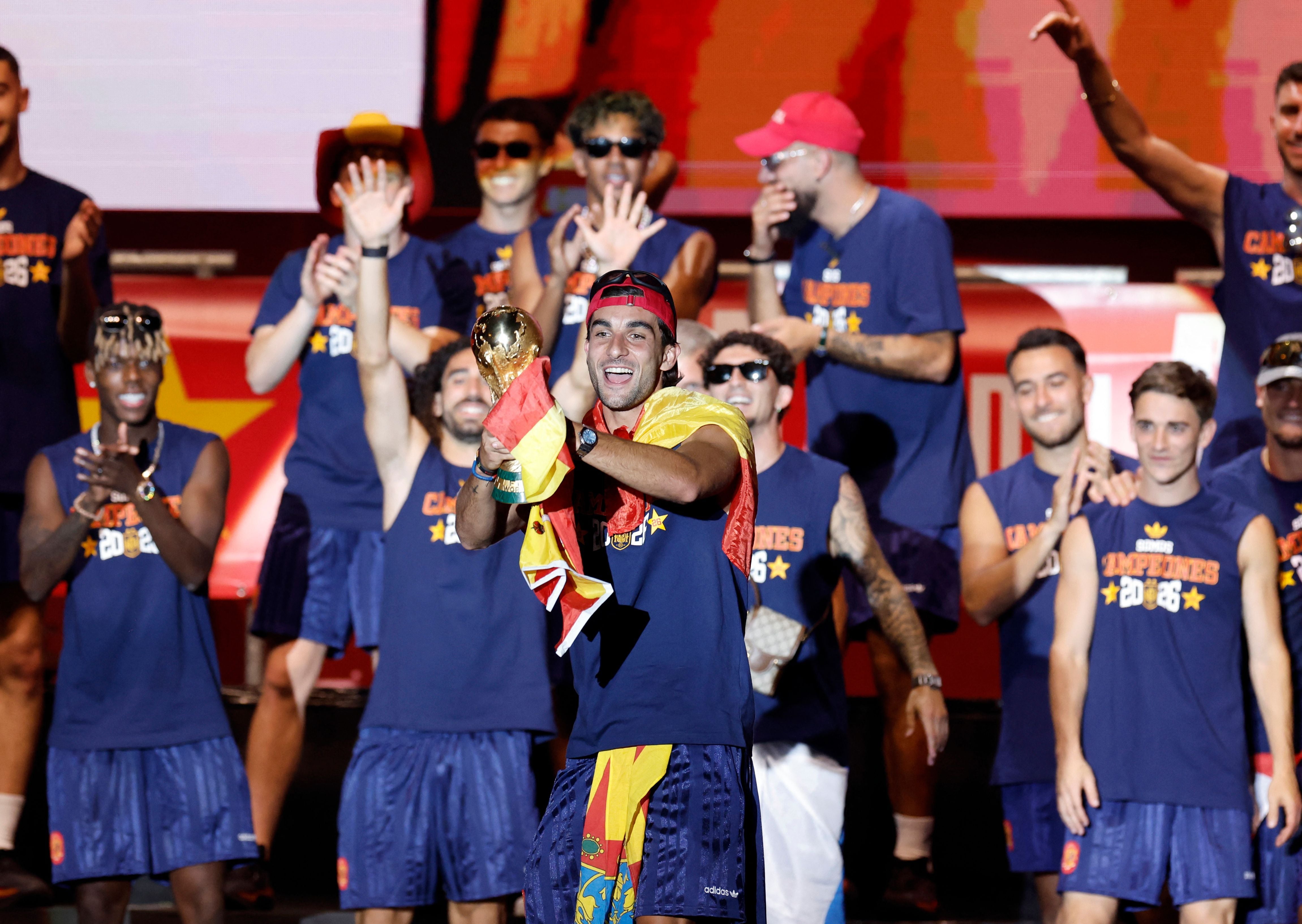 Spaniens Fußballspieler feiern auf einer Bühne, ein Spieler hält eine Trophäe und trägt eine Flagge.