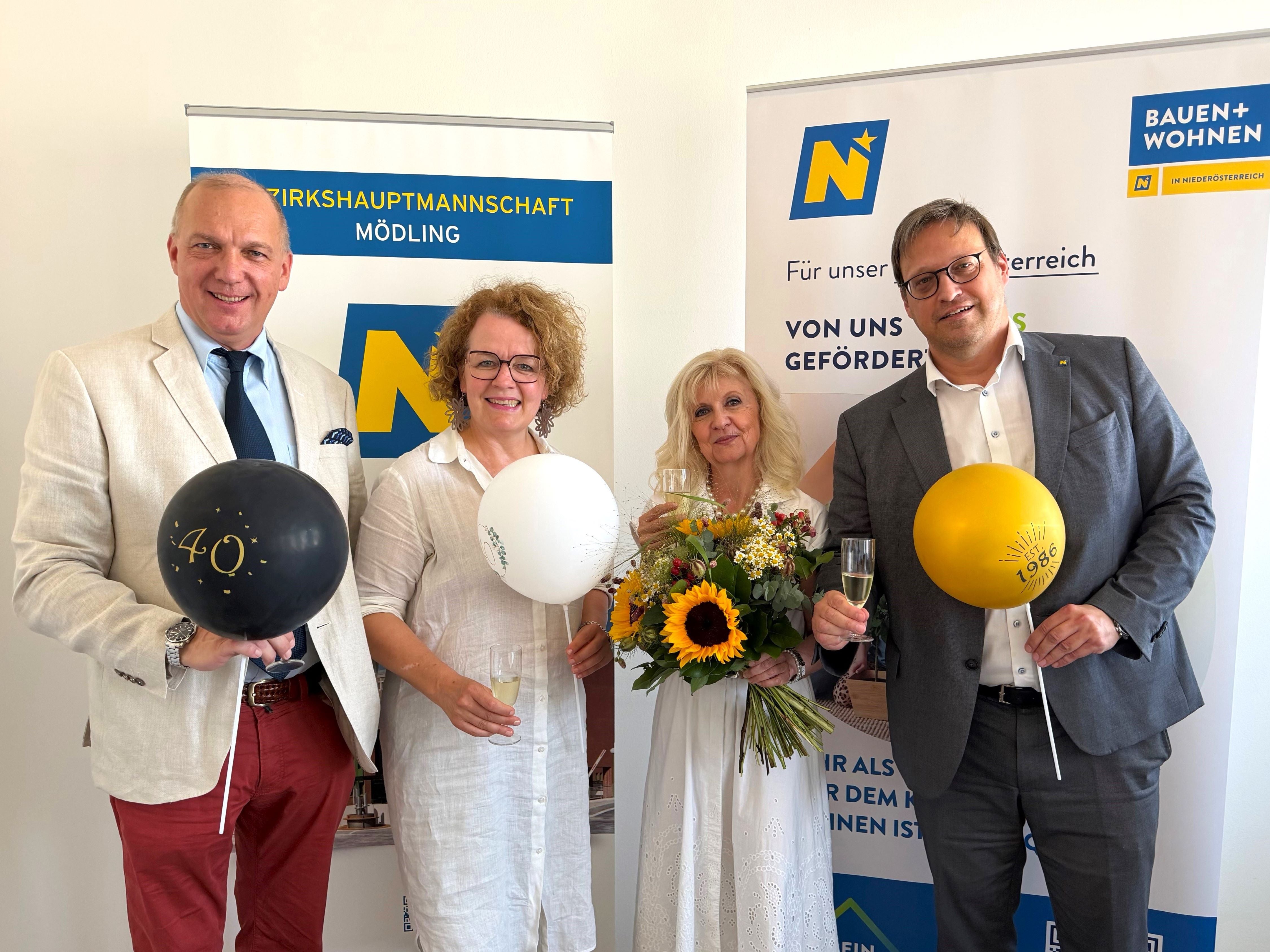 Lächelnd mit Luftballons und Blumenstrauß vor Info-Bannern.