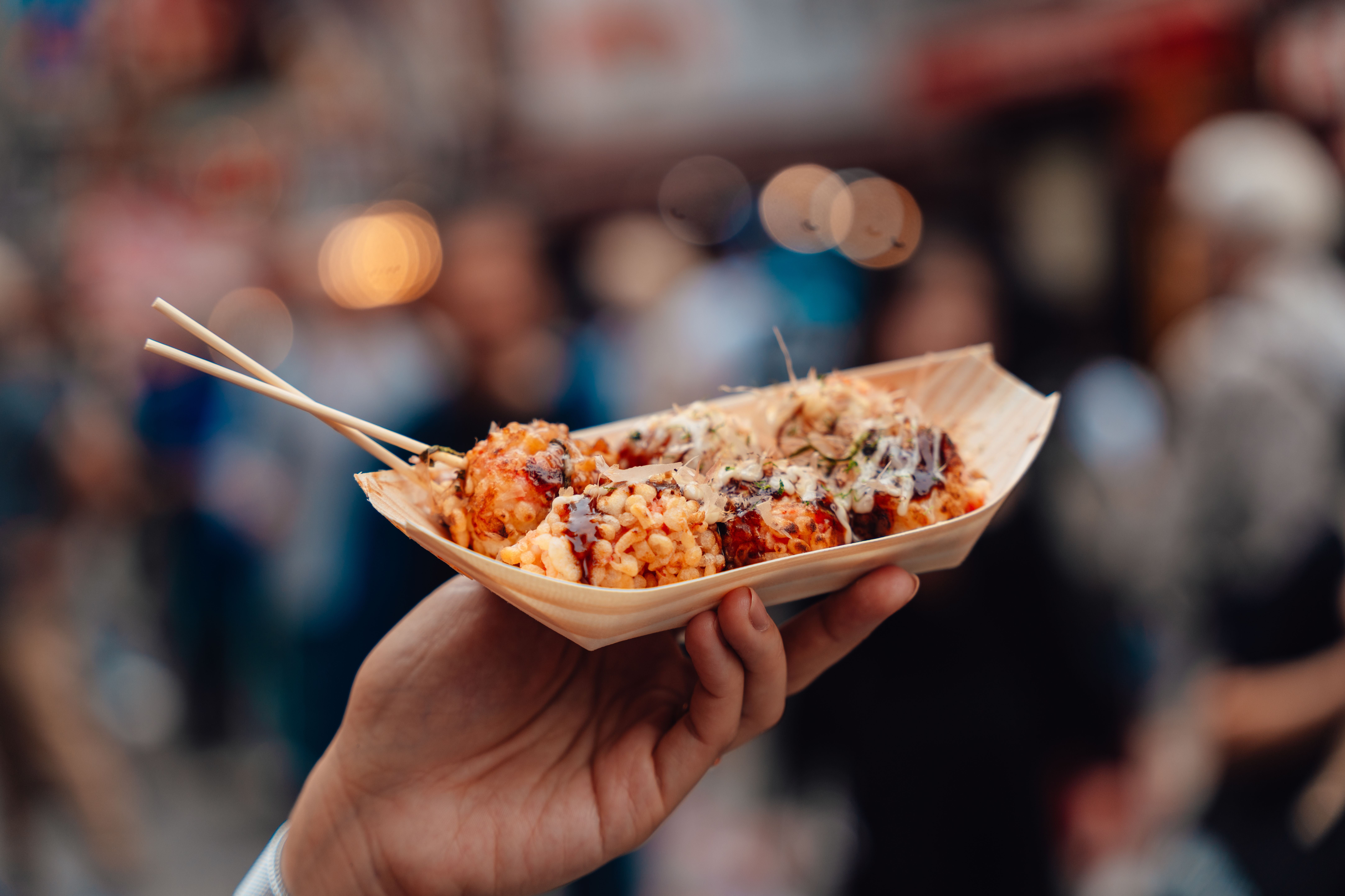 Eine Hand hält Takoyaki in einem Holzschälchen mit Stäbchen auf einer belebten Straße.
