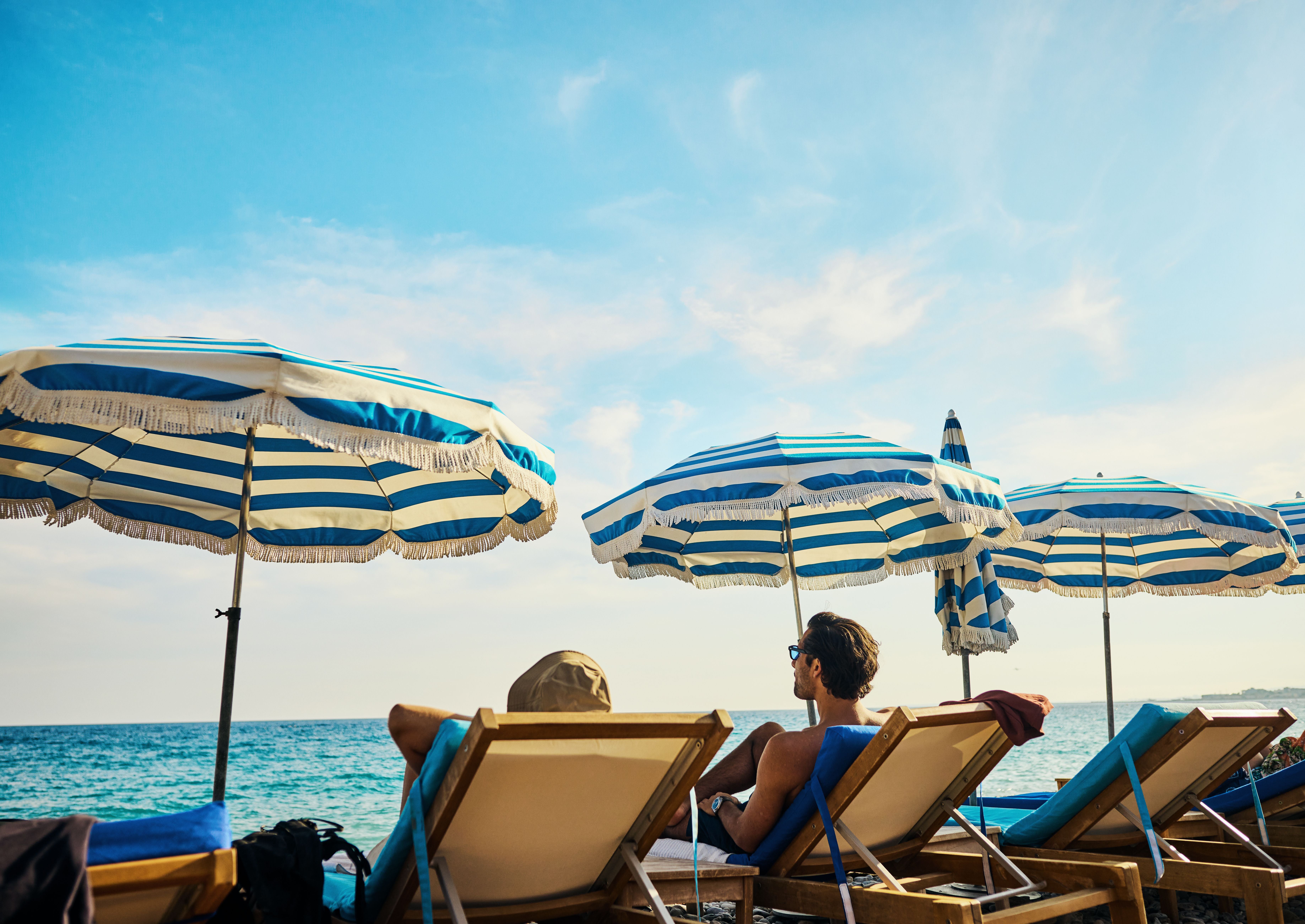 Zwei Personen entspannen auf Liegen unter gestreiften Sonnenschirmen am Strand mit Meerblick.
