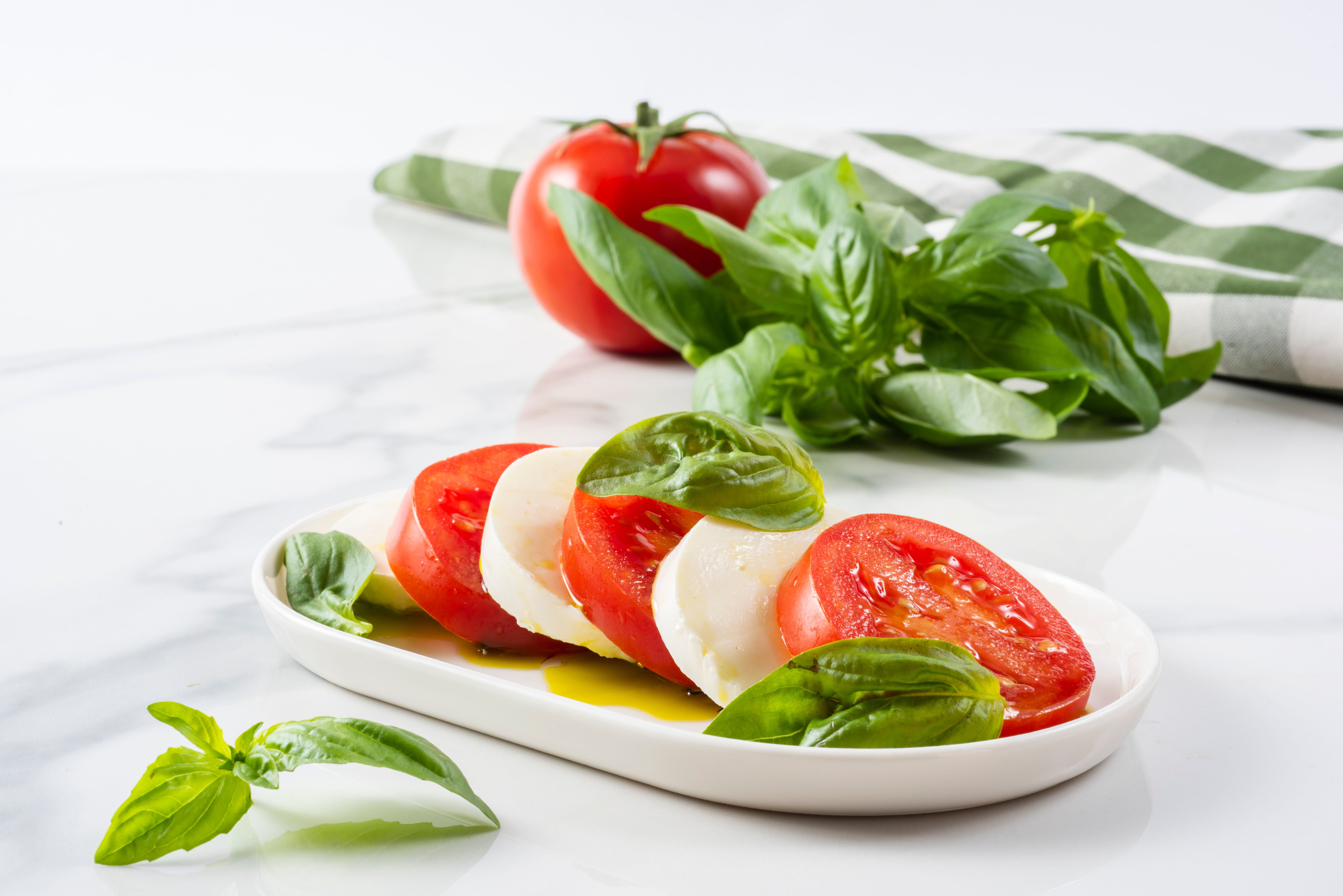 Caprese-Salat mit Tomaten, Mozzarella und Basilikum auf weißem Teller, im Hintergrund frische Kräuter.