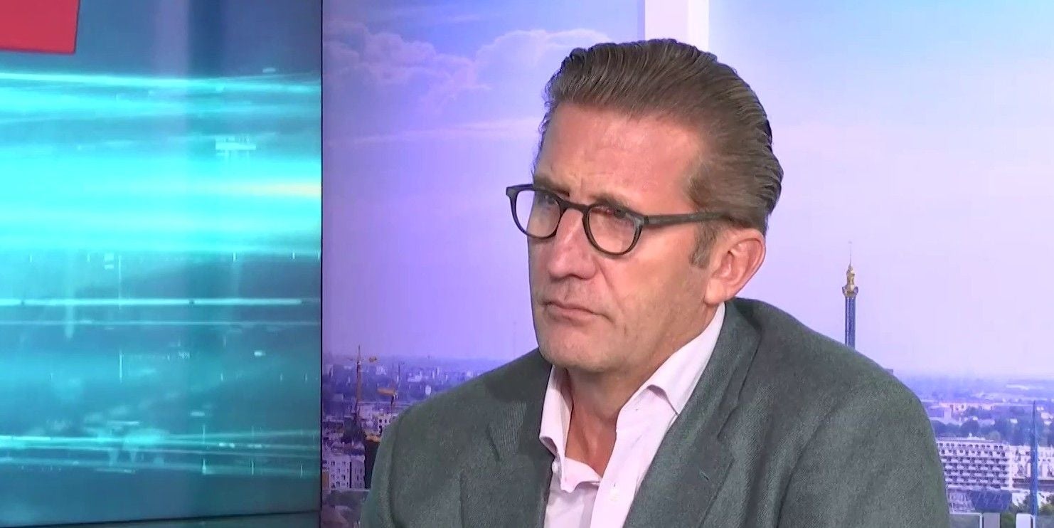 Ein Mann mit Brille und grauem Anzug sitzt vor einem Stadtpanorama im Fernsehstudio.