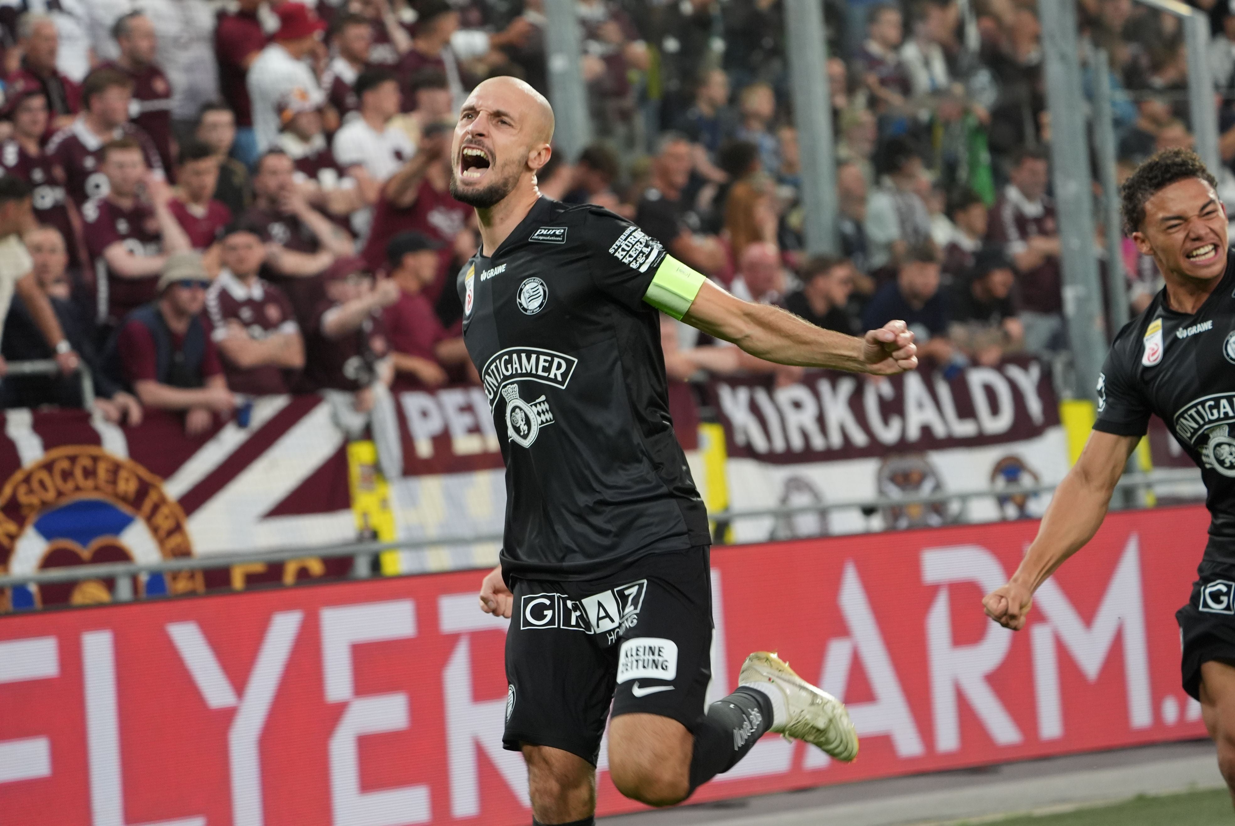 Ein glatzköpfiger Fußballspieler im schwarzen Sturm-Graz-Trikot mit Kapitänsbinde am Arm brüllt voller Freude mit weit geöffnetem Mund. Er steht auf dem Spielfeld nahe der Bande, während im Hintergrund Fans auf den Rängen zu sehen sind.