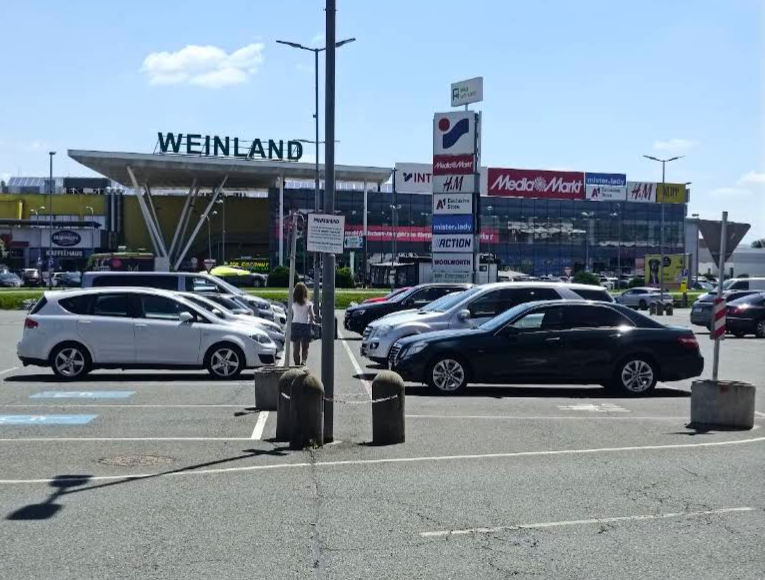 Parkplatz vor einem Einkaufszentrum mit mehreren Autos und einer Person, darüber das Schild "Weinland".
