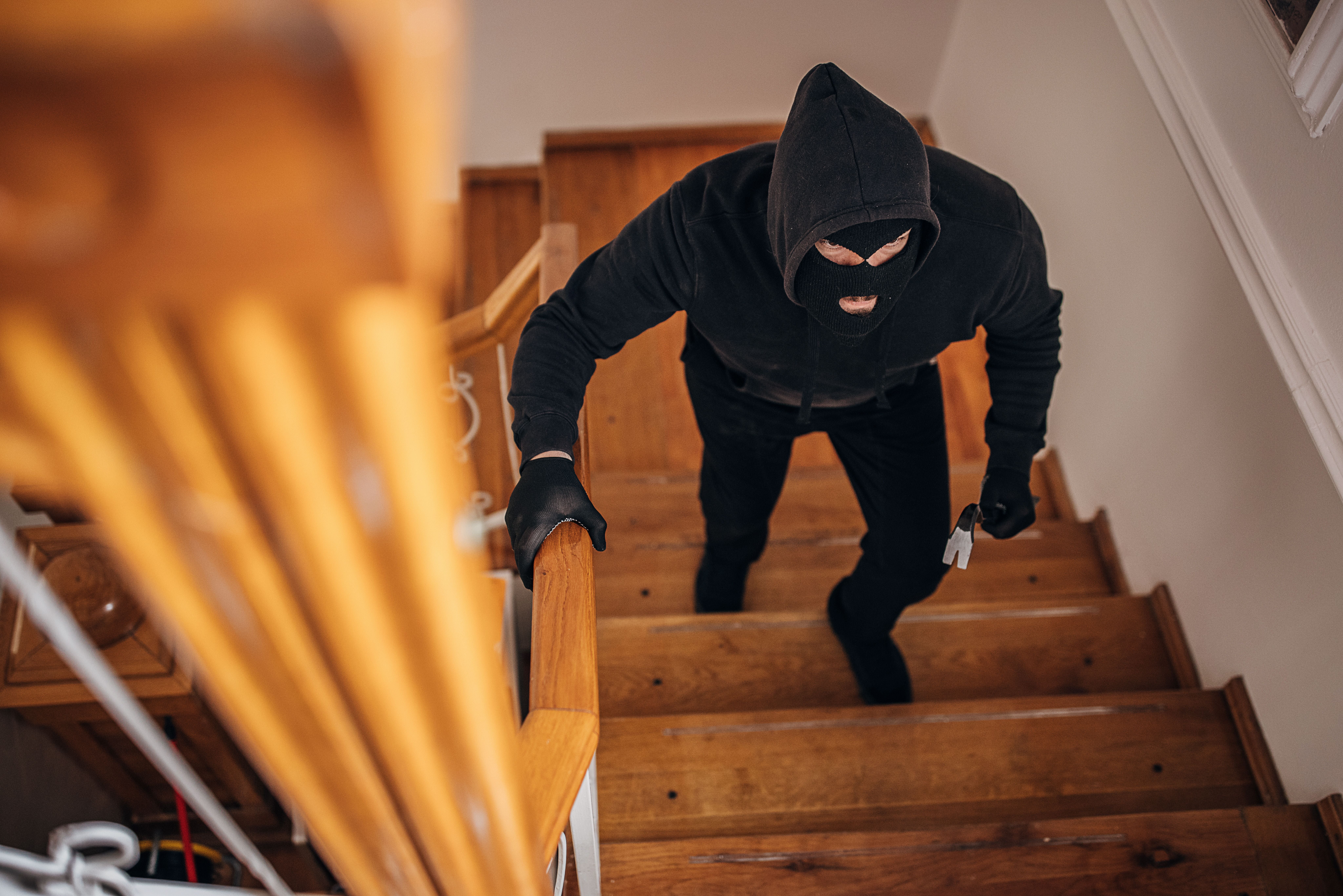 Maskierter Einbrecher in schwarzer Kleidung läuft mit Brecheisen eine Treppe hoch.