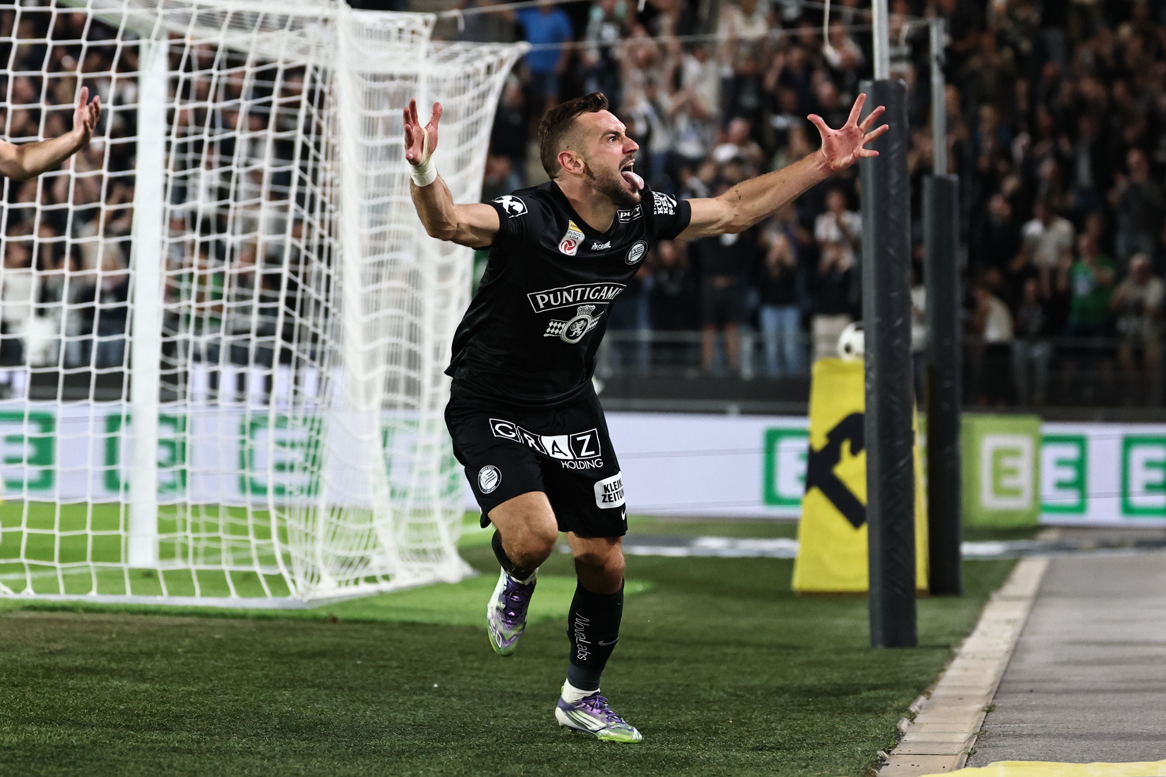 Ein Fußballspieler in einem schwarzen Trikot mit kurzen Ärmeln rennt jubelnd mit ausgebreiteten Arme und geöffnetem Mund nach rechts. Er befindet sich auf einem grünen Spielfeld nahe der Torauslinie, direkt vor einem weißen Tornetz. Im Hintergrund stehen Fans auf den Rängen.