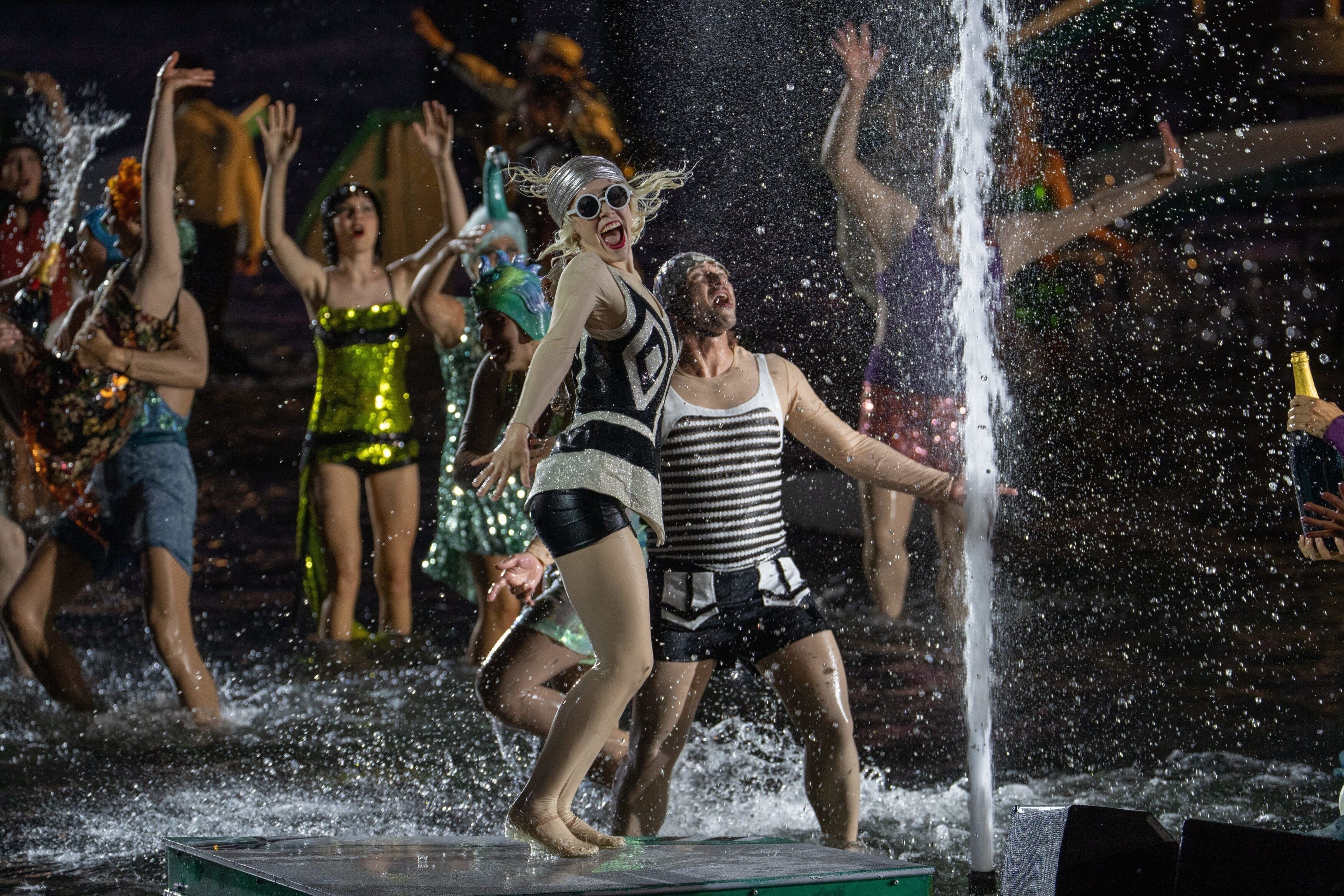 Enthusiastische Tänzer in 20er-Jahre-Kostümen feiern auf einer nassen Bühne mit Wasserfontäne.
