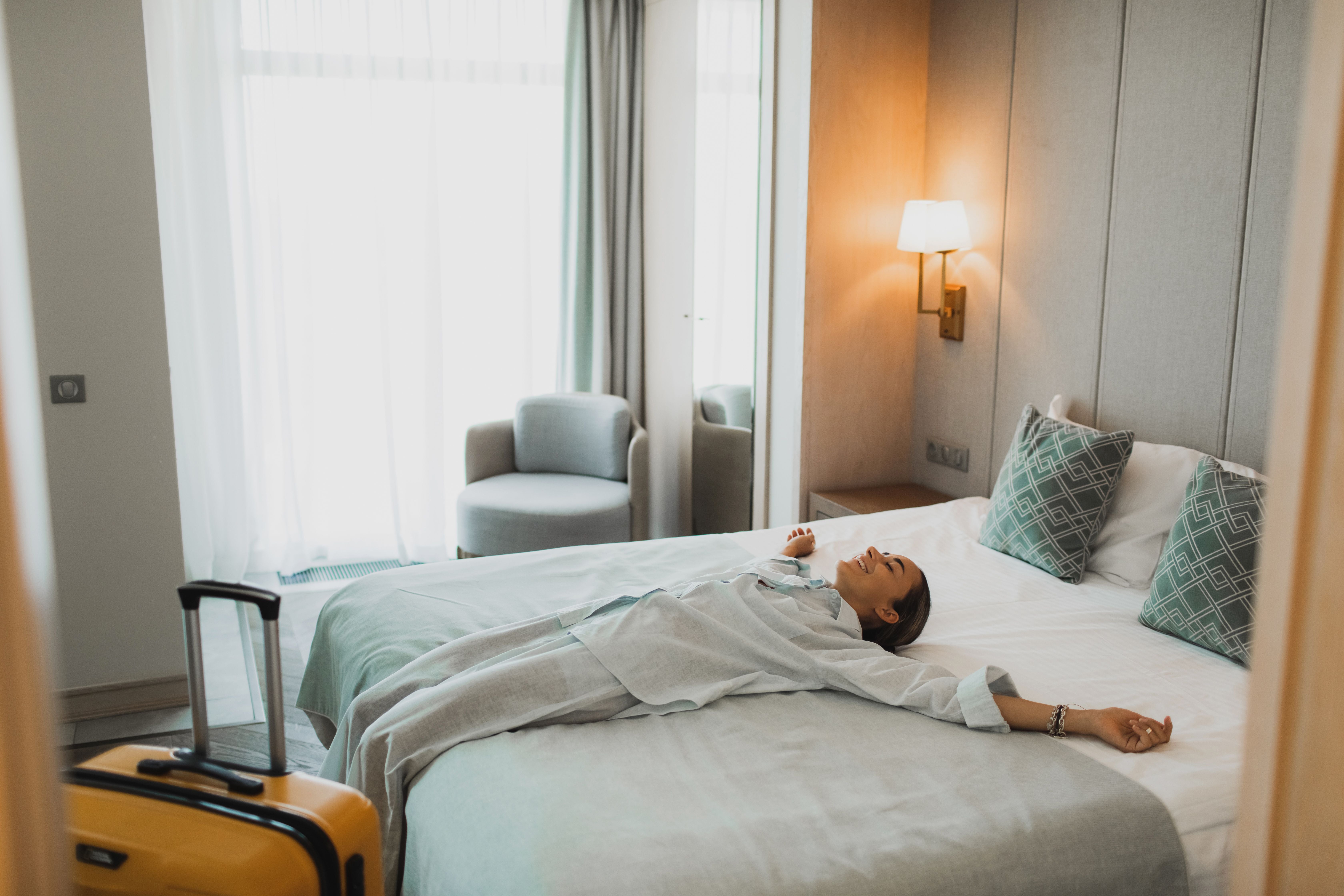 Frau liegt entspannt lachend auf dem Bett in einem Hotelzimmer, gelber Koffer im Vordergrund.