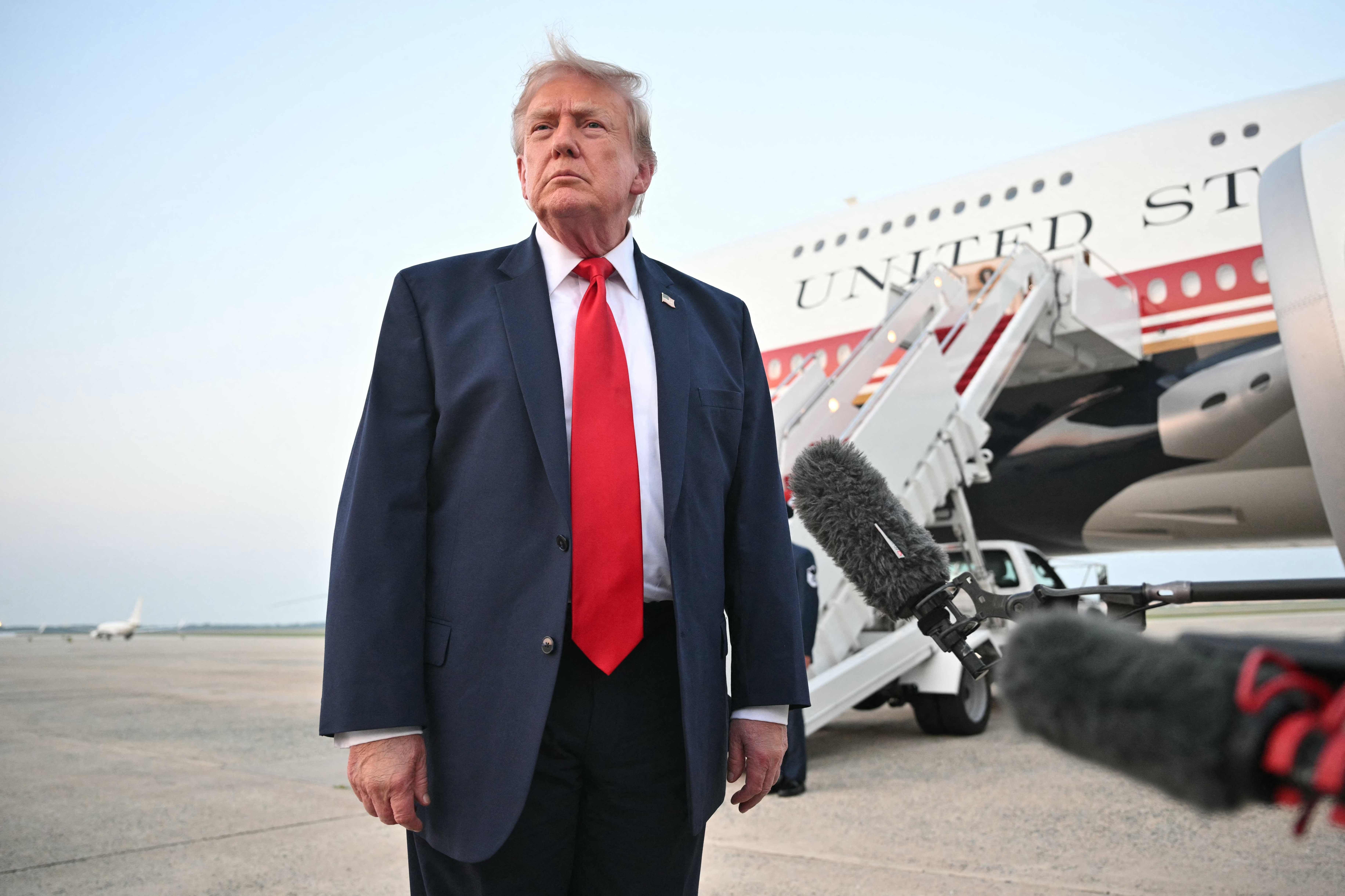 Ein Mann im Anzug steht vor einem Flugzeug und spricht zu Mikrofonen auf dem Rollfeld.
