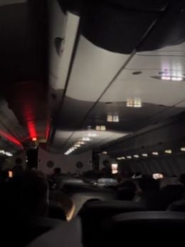 Dunkle Flugzeugkabine mit Passagieren und reflektierten Lichtern an der Decke.