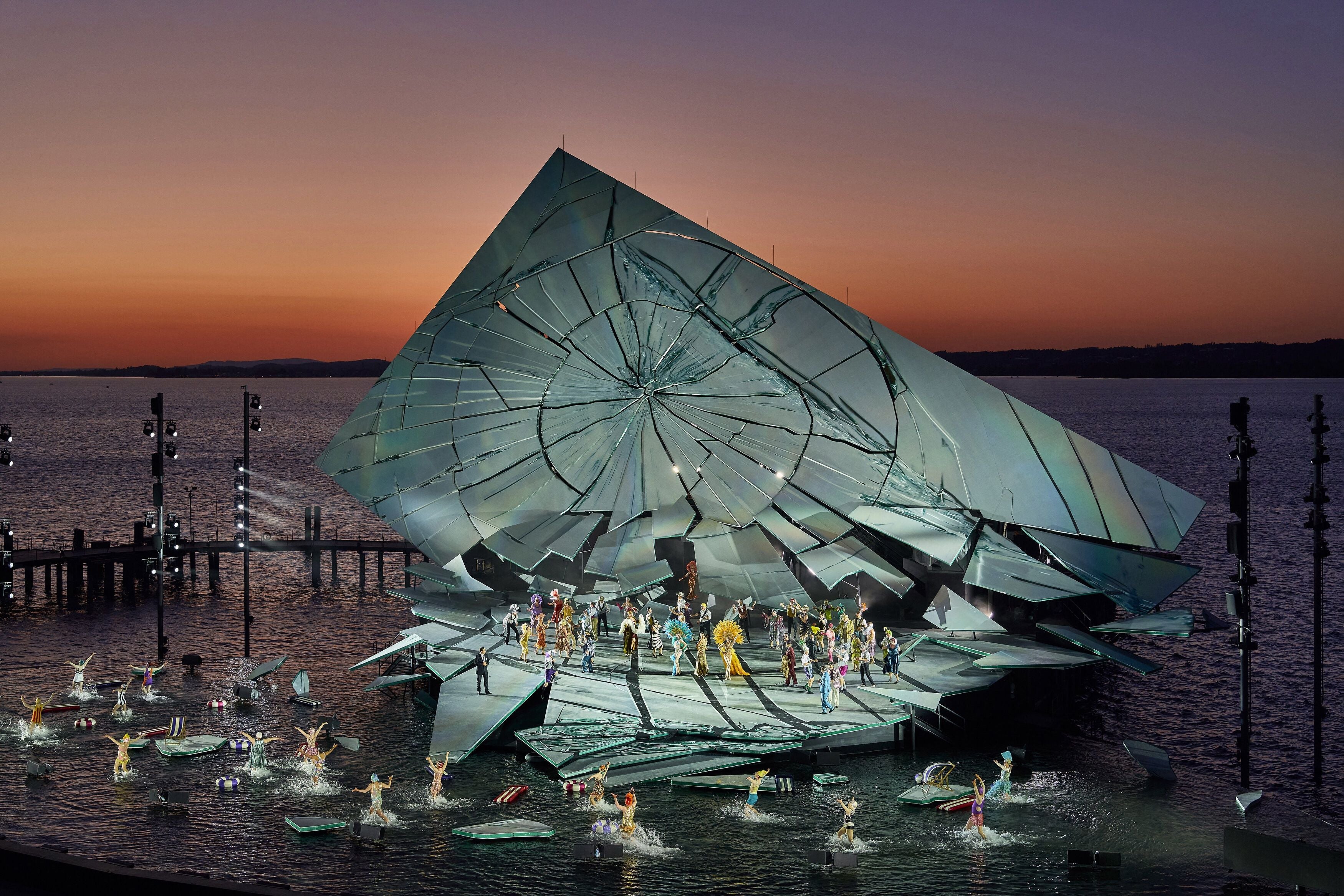 Open-Air-Opernbühne auf dem Wasser bei Sonnenuntergang, mit großem zerbrochenem Spiegel-Set und Darstellern.