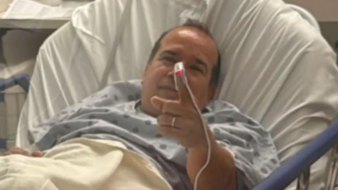 Ein Mann liegt im Krankenhausbett und zeigt mit einem Finger, an dem ein Pulsoximeter hängt.