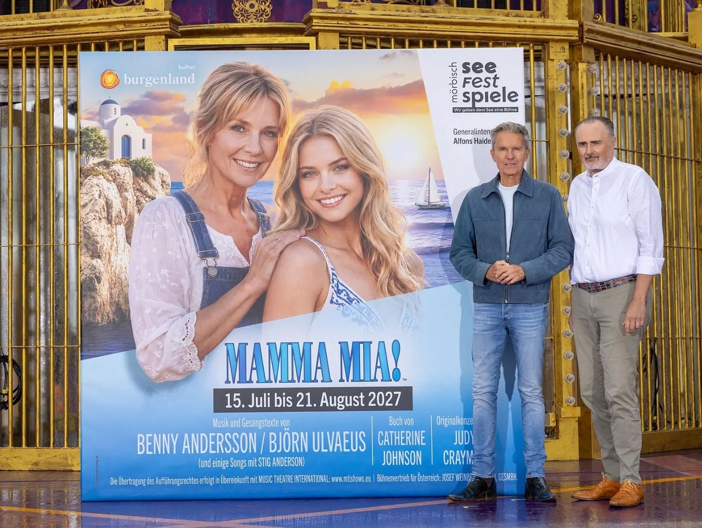 Zwei Männer stehen neben einem großen Mamma Mia!-Werbeplakat vor einem goldenen Zaun.