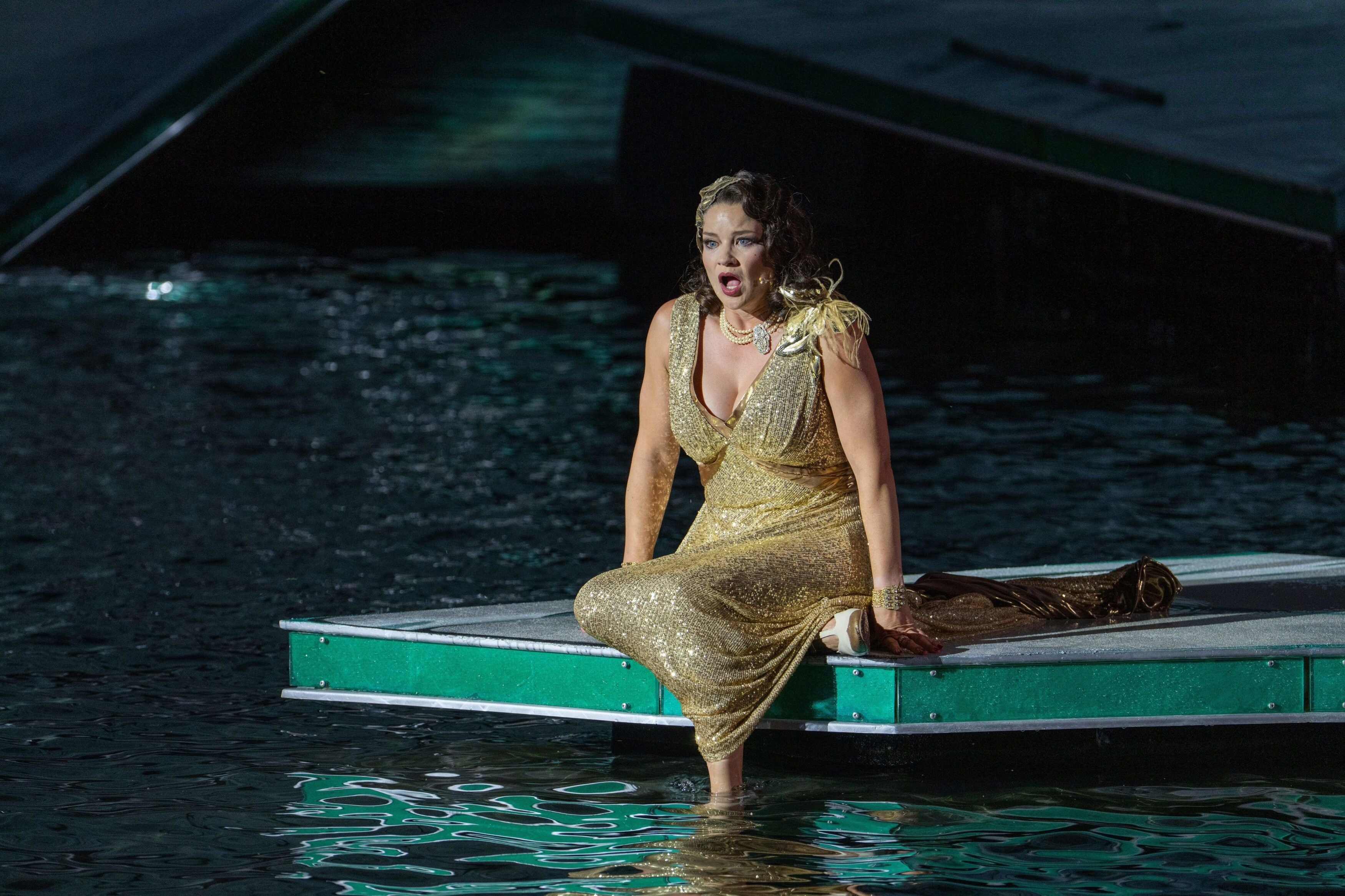 Eine Frau in goldenem Kleid sitzt auf einer Bühne am Wasser und singt leidenschaftlich.