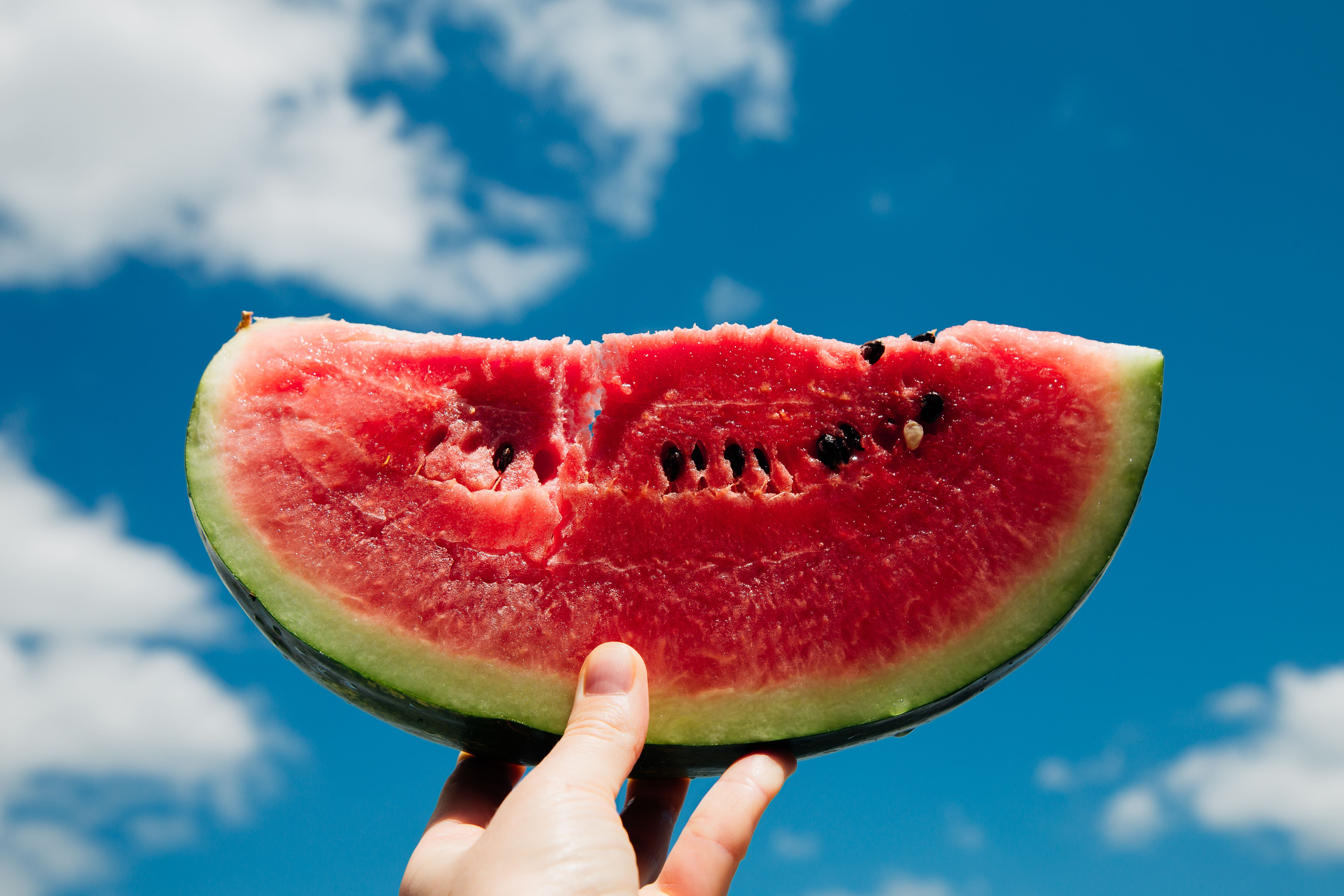 Eine Hand hält ein großes Stück Wassermelone vor blauem Himmel mit Wolken.