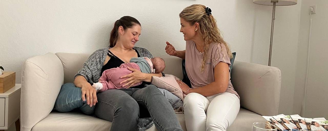 Zwei Frauen sitzen auf dem Sofa, eine stillt ein Baby, die andere spricht mit ihr.