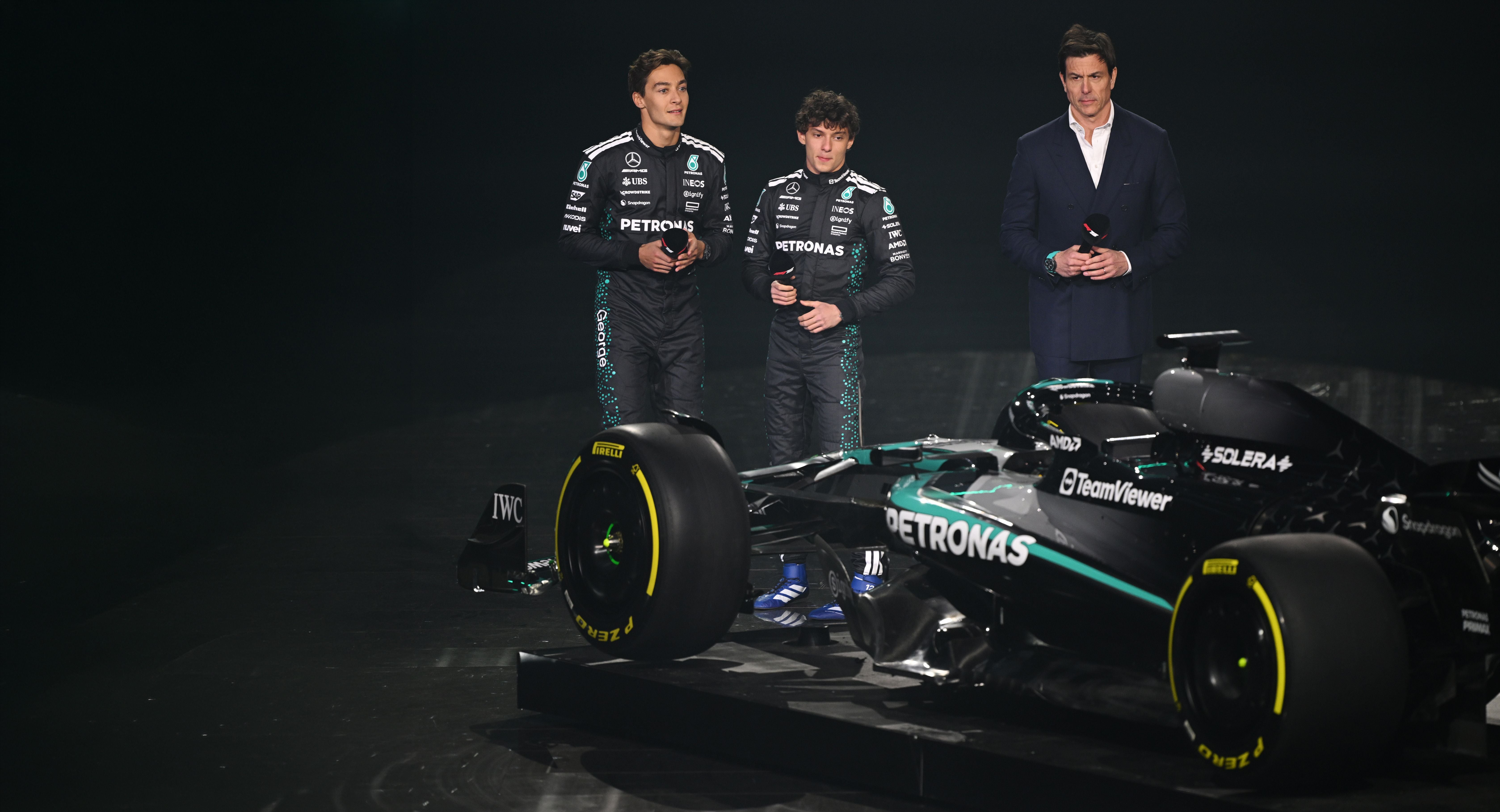 George Russell, Kimi Antonelli und Toto Wolff stehen nebeneinander auf einer dunklen Bühne hinter einem schwarzen Mercedes-Formel-1-Rennwagen.