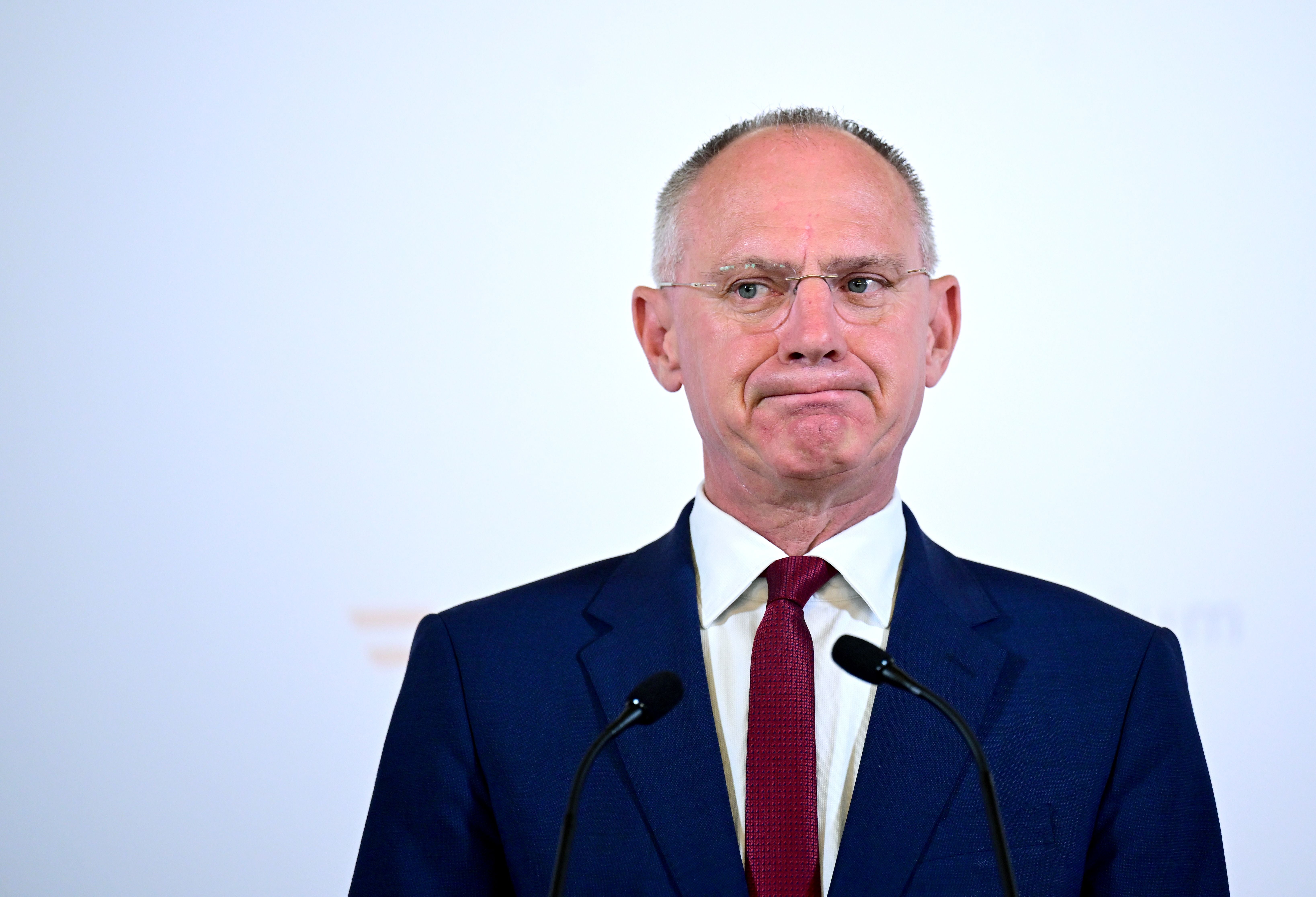 Ein Mann im Anzug und mit Brille steht vor Mikrofonen vor weißem Hintergrund.
