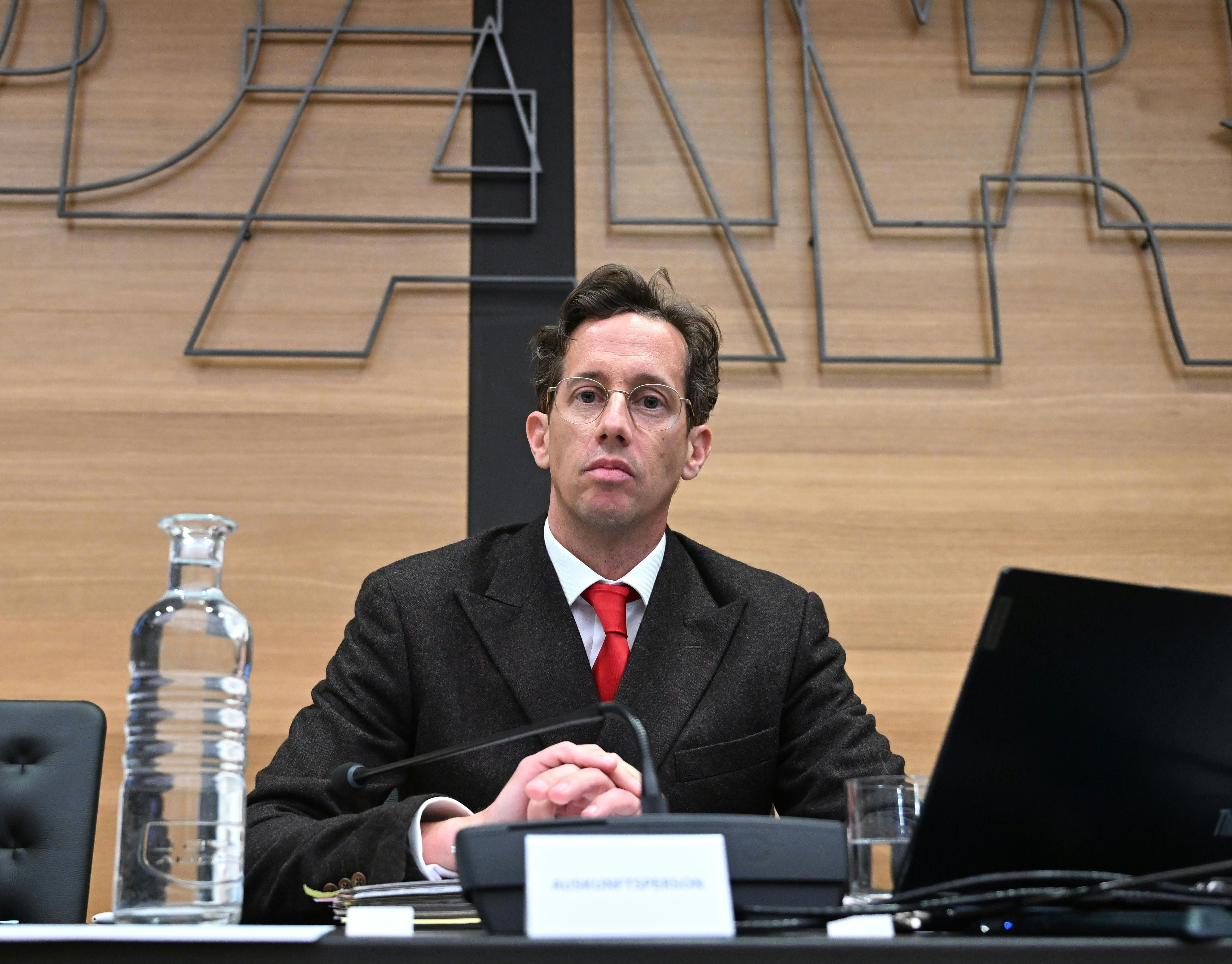Ein Mann im Anzug mit roter Krawatte sitzt bei einer Sitzung im Parlament vor Laptop und Mikrofon.