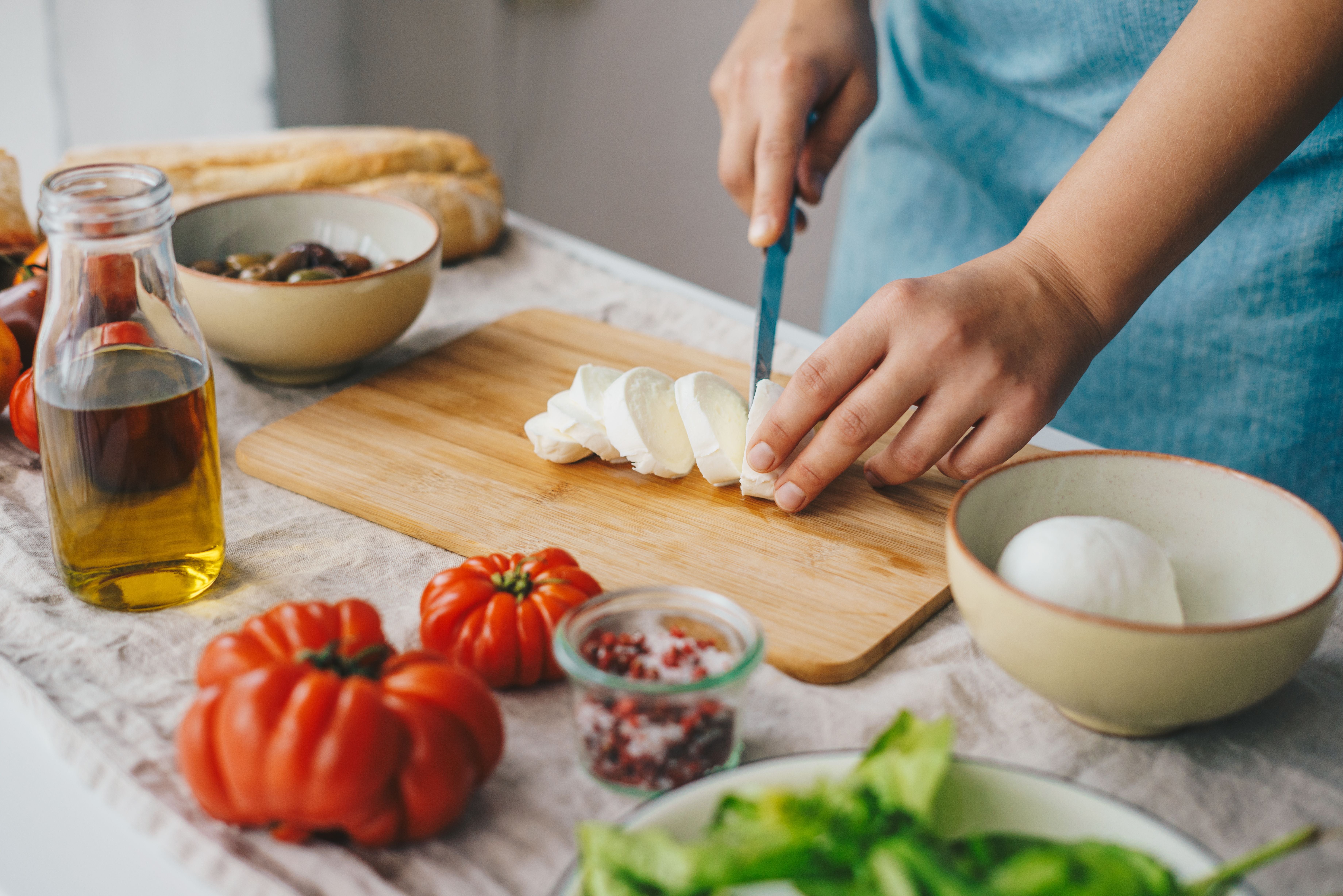 Frau schneidet Mozzarella für Caprese-Salat mit Tomaten, Basilikum und Olivenöl.