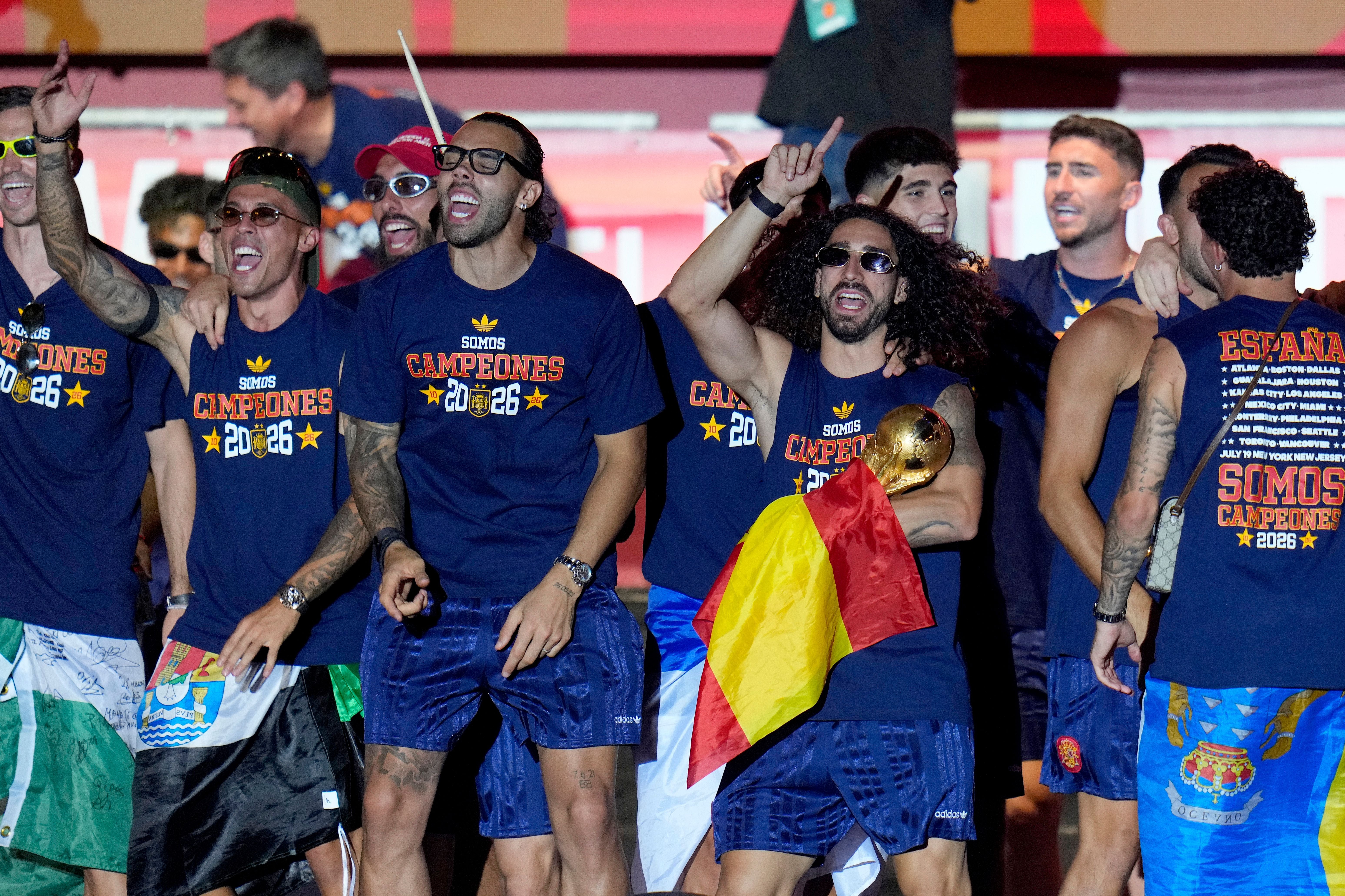 Spanische Fußballspieler feiern auf einer Bühne mit T-Shirts „SOMOS CAMPEONES 2026“ und einer Trophäe.