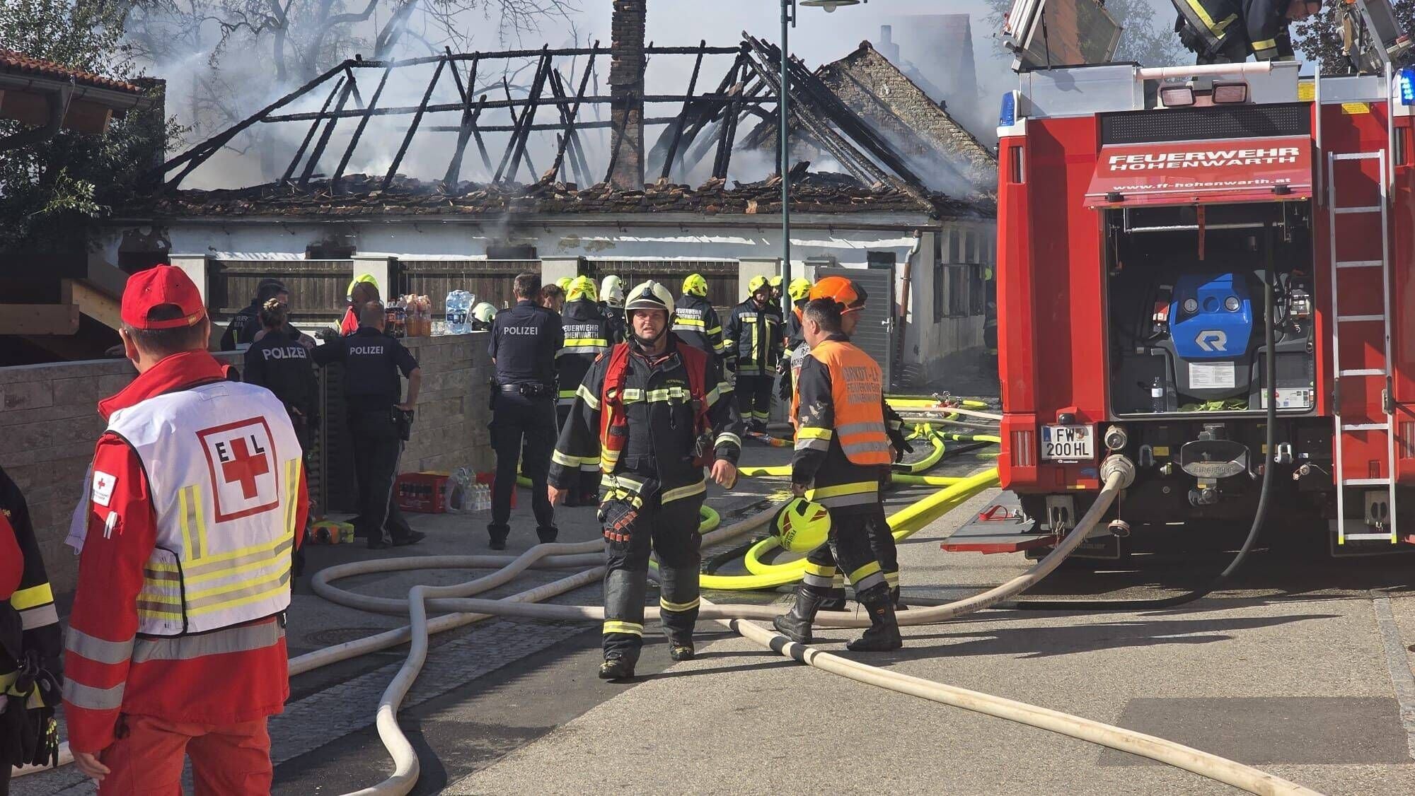 Feuerwehr und Polizei bekämpfen Brand in Einfamilienhaus mit zerstörtem Dach, Rauch steigt auf.