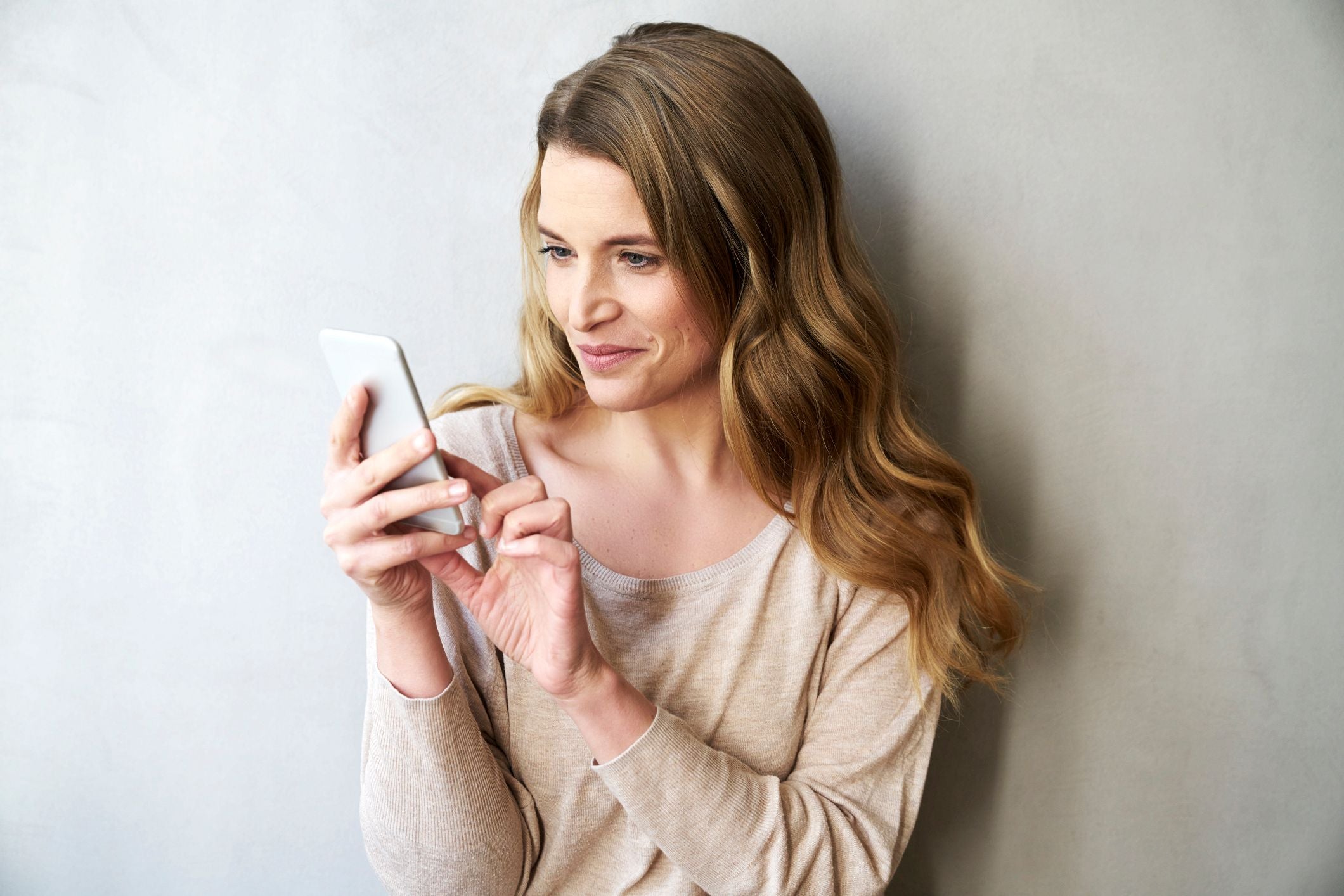 Junge Frau mit langen Haaren lächelt und tippt auf ihr Smartphone vor hellgrauer Wand.