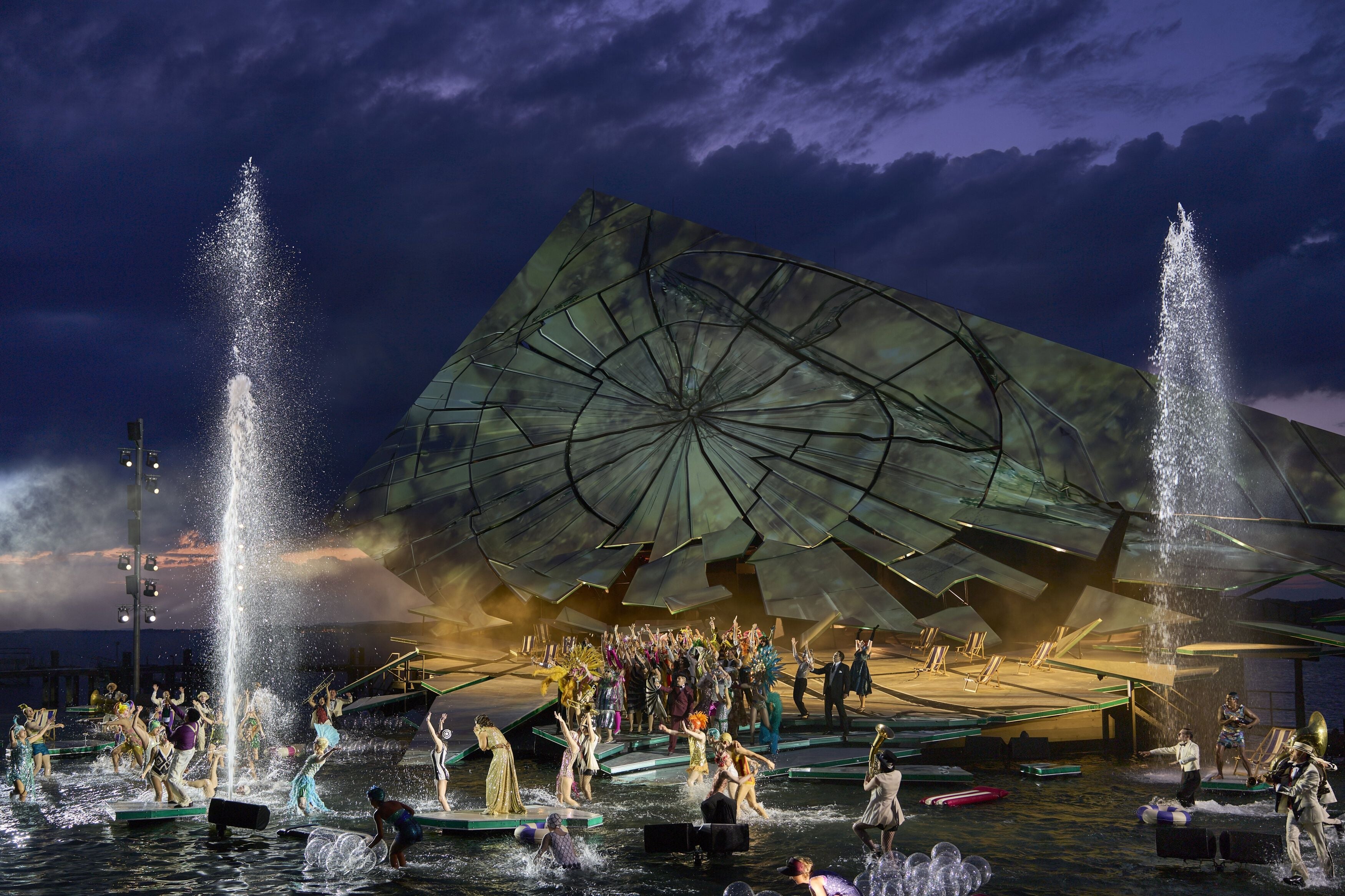 Bühnenbild der Bregenzer Festspiele mit vielen Darstellern vor riesiger Scheibe und Wasserfontänen.